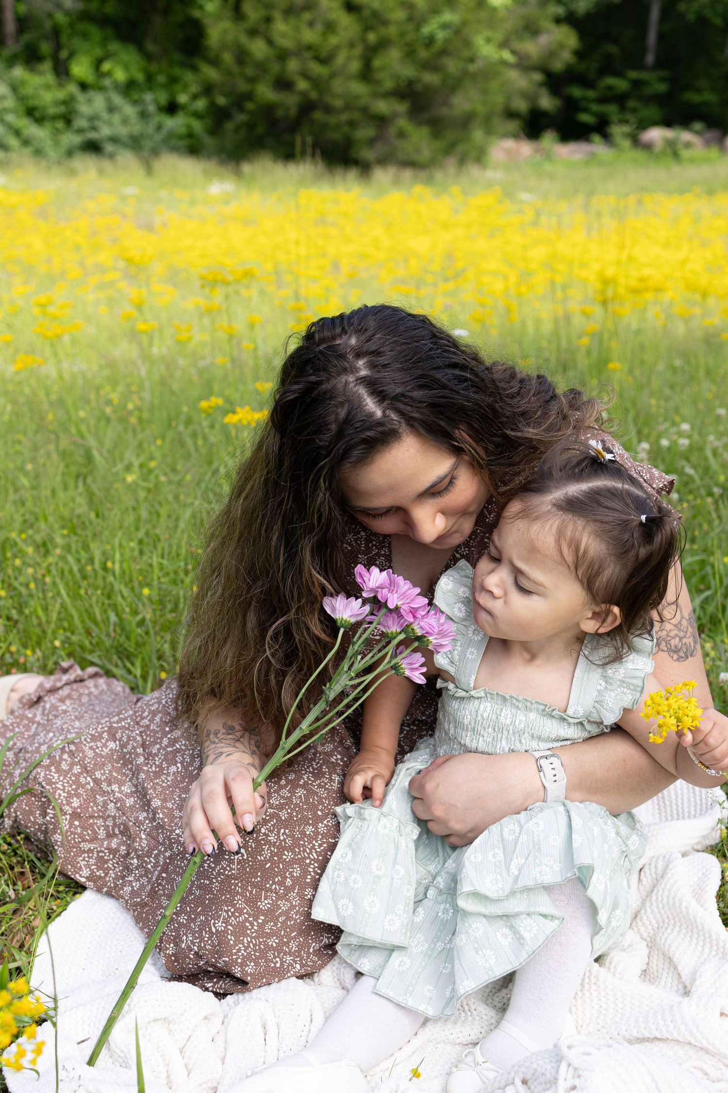 Mother kissing toddler daughter holding pink wildflowers while sitting on blanket in sunny yellow flower field.
