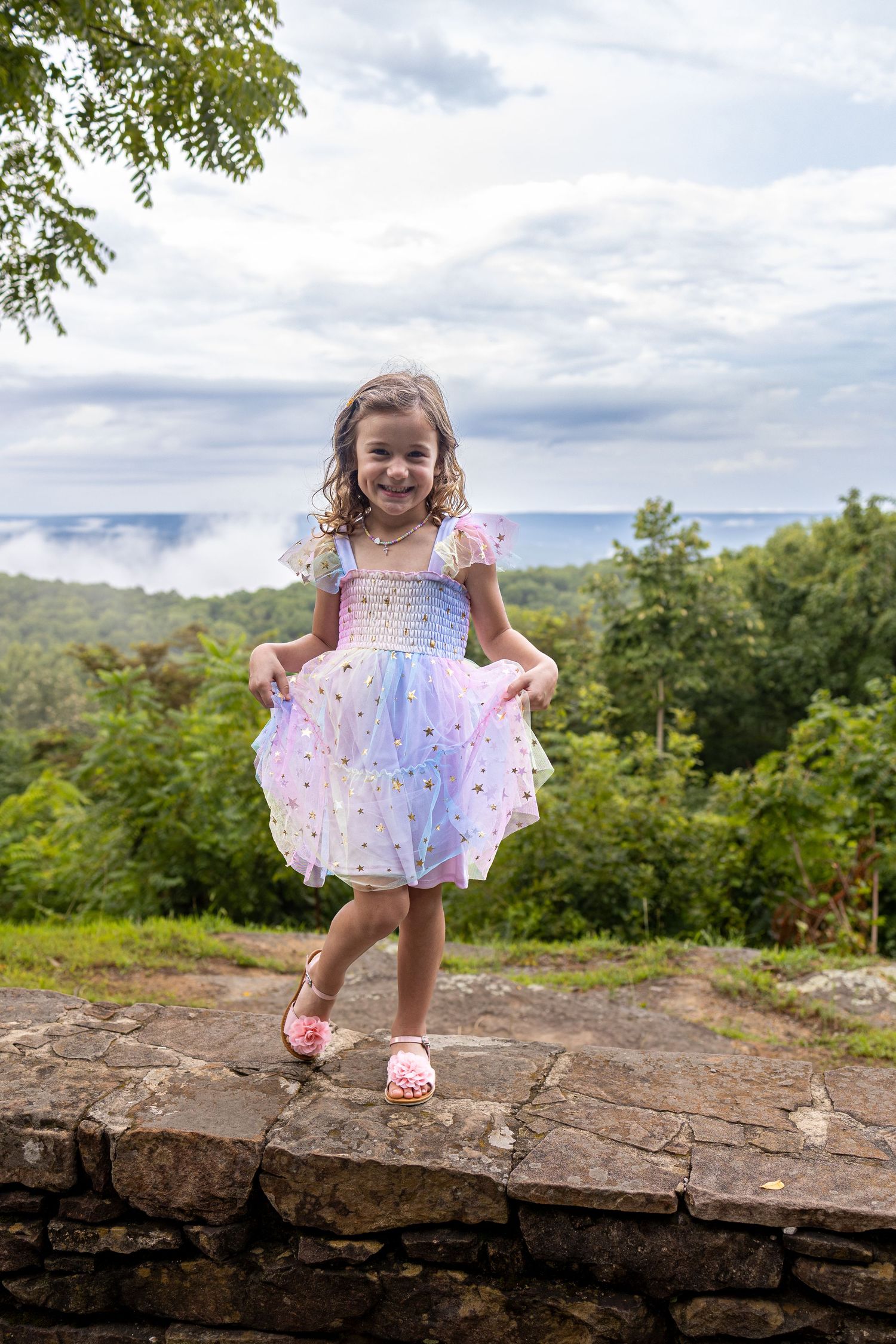 A young girl in a colorful tutu dress smiles while standing on a stone wall with lush green trees behind her.