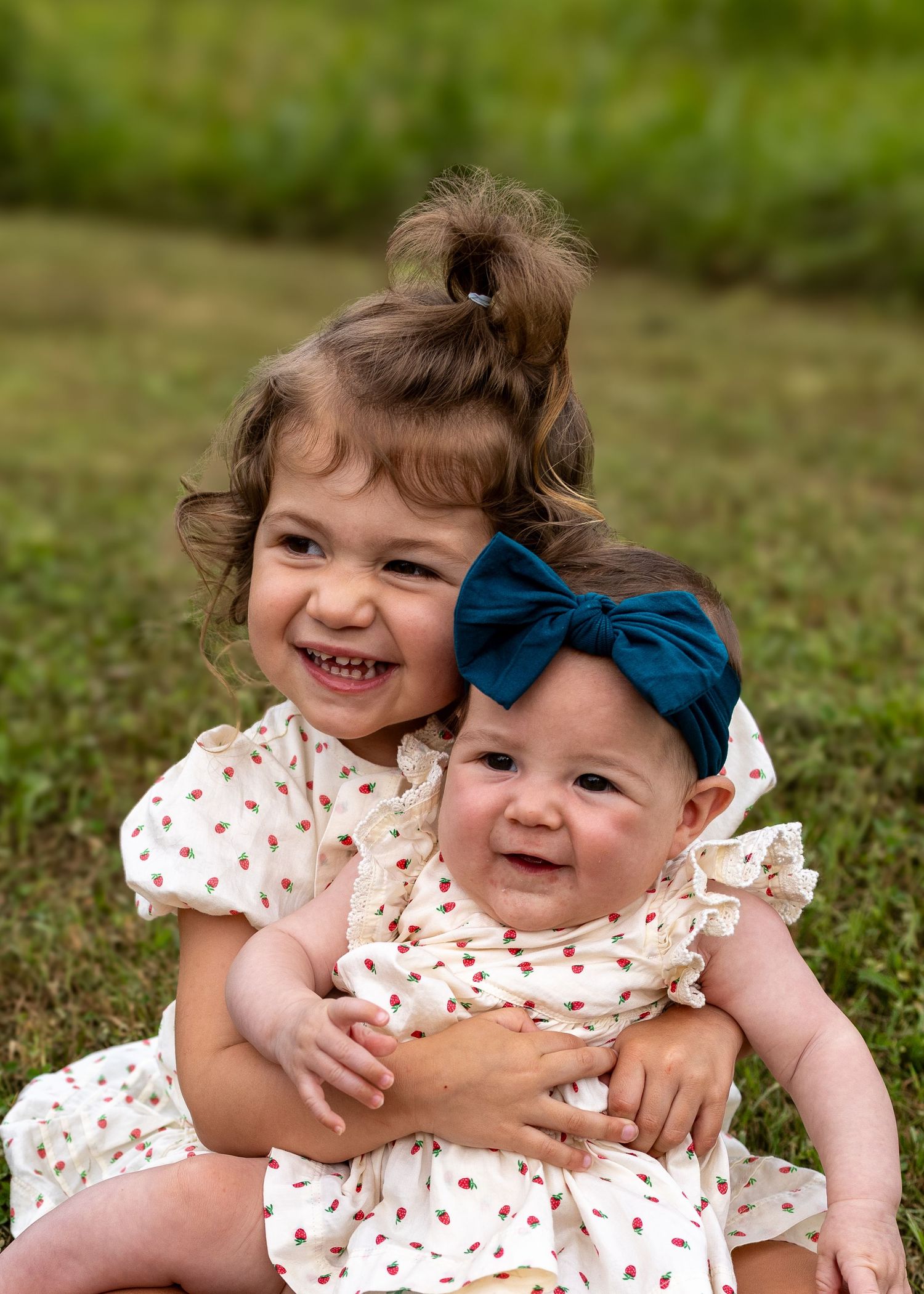 Two smiling sisters in matching floral dresses sit together on green grass, older girl hugging baby with navy bow headband.