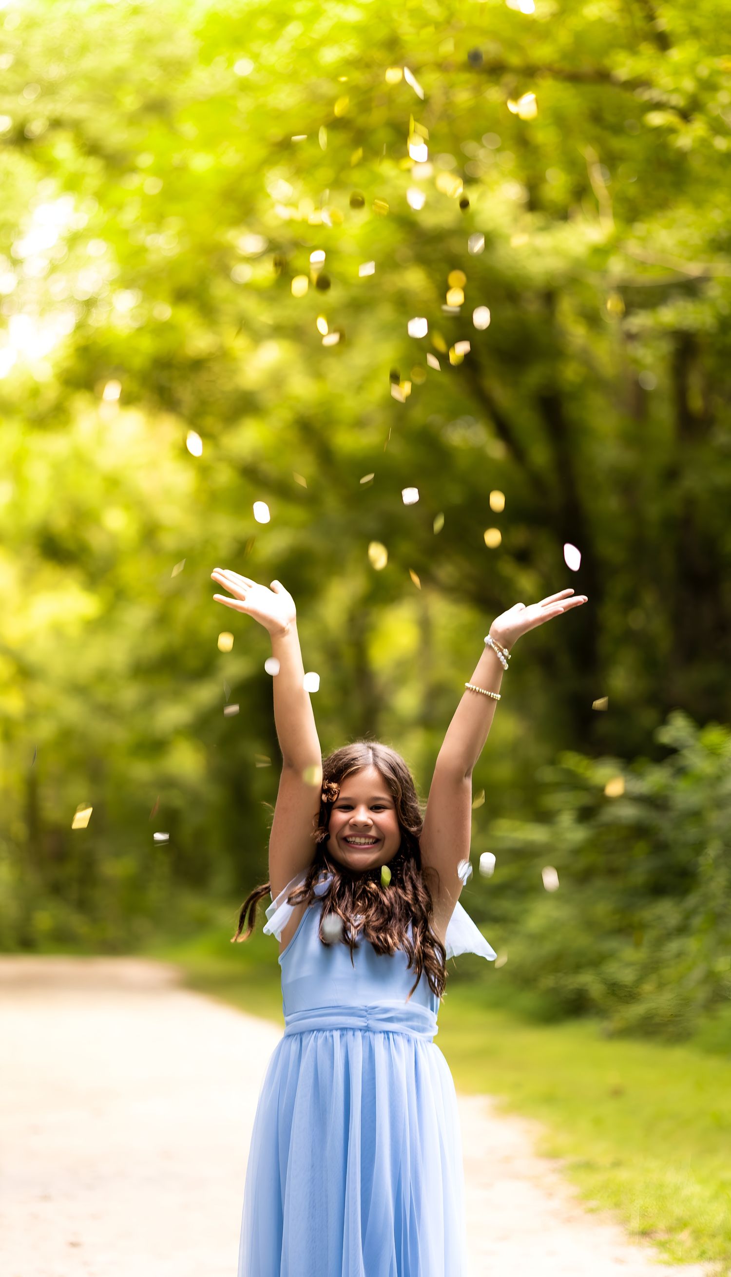 A joyful girl in a blue dress tosses white flower petals into the air on a tree-lined path.