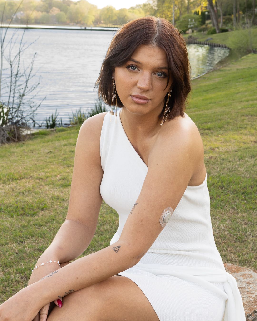 Young woman in white one-shoulder dress sitting by a lake on green grass, wearing gold earrings and bracelet.