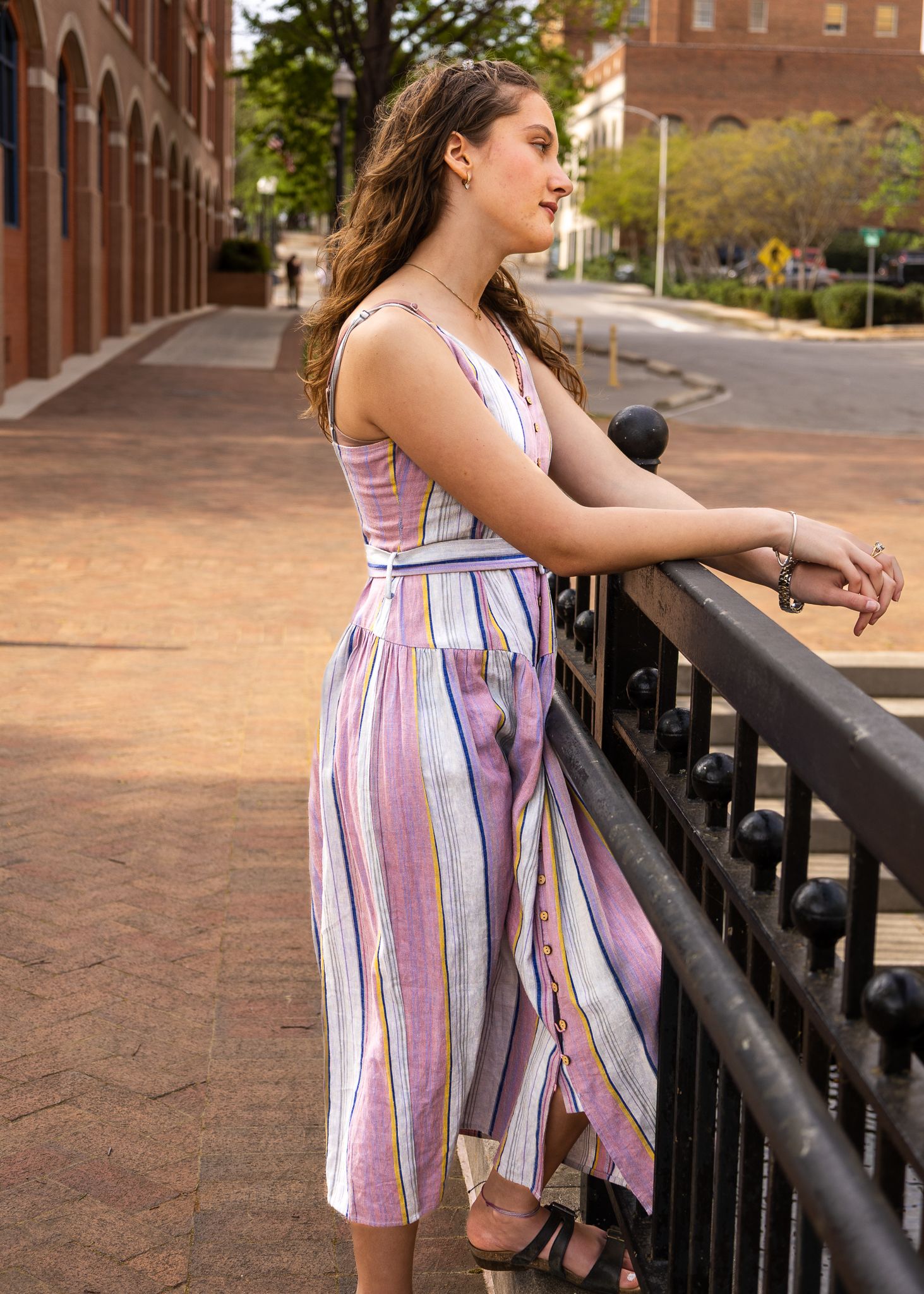 Young woman in colorful striped midi dress leaning on black iron railing on a sunny urban street.