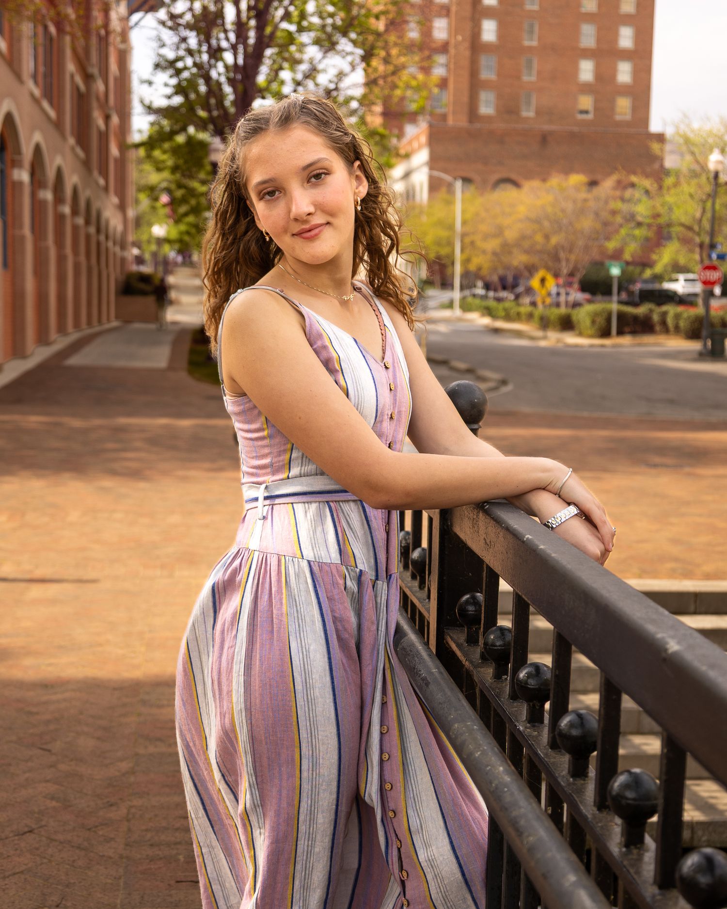 Young woman in colorful striped jumpsuit leaning on iron railing in urban brick streetscape setting.