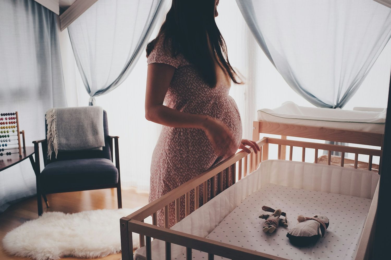 Pregnant woman in lace dress standing by a wooden baby crib in a softly lit nursery room.