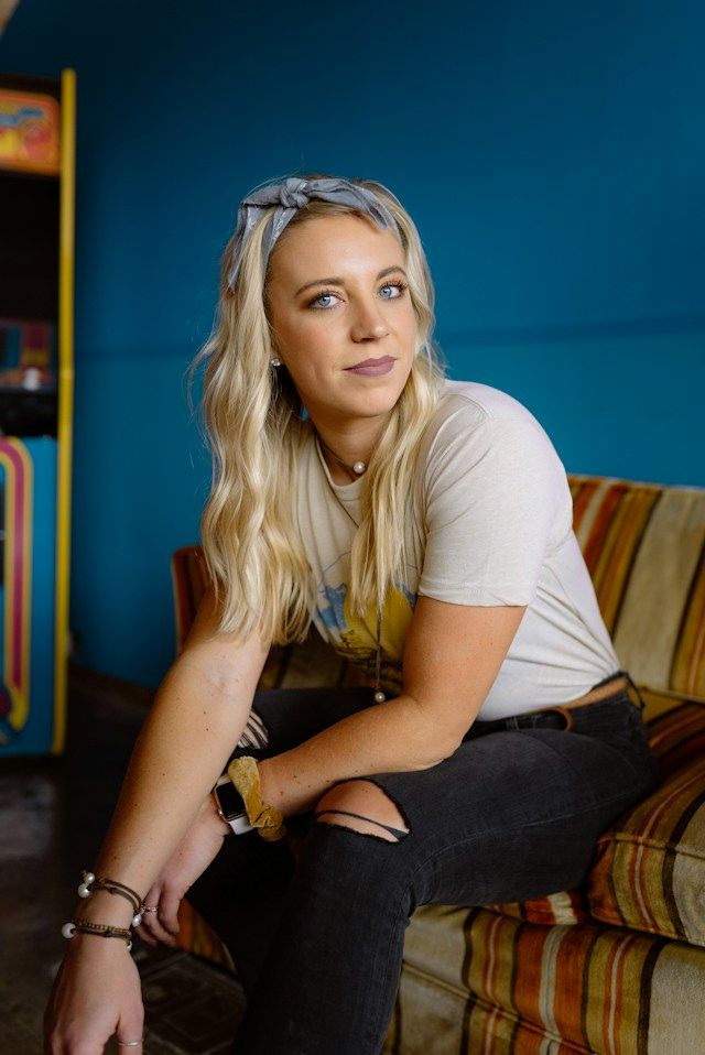Blonde woman in white t-shirt and ripped jeans sits on striped couch in colorful blue room.