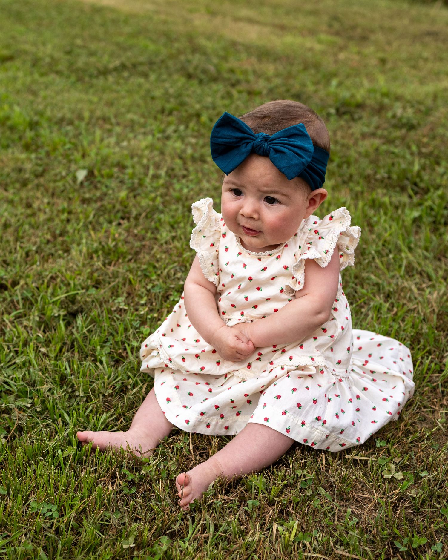 Smiling baby girl in white floral dress and teal bow headband sitting on green grass outdoors.