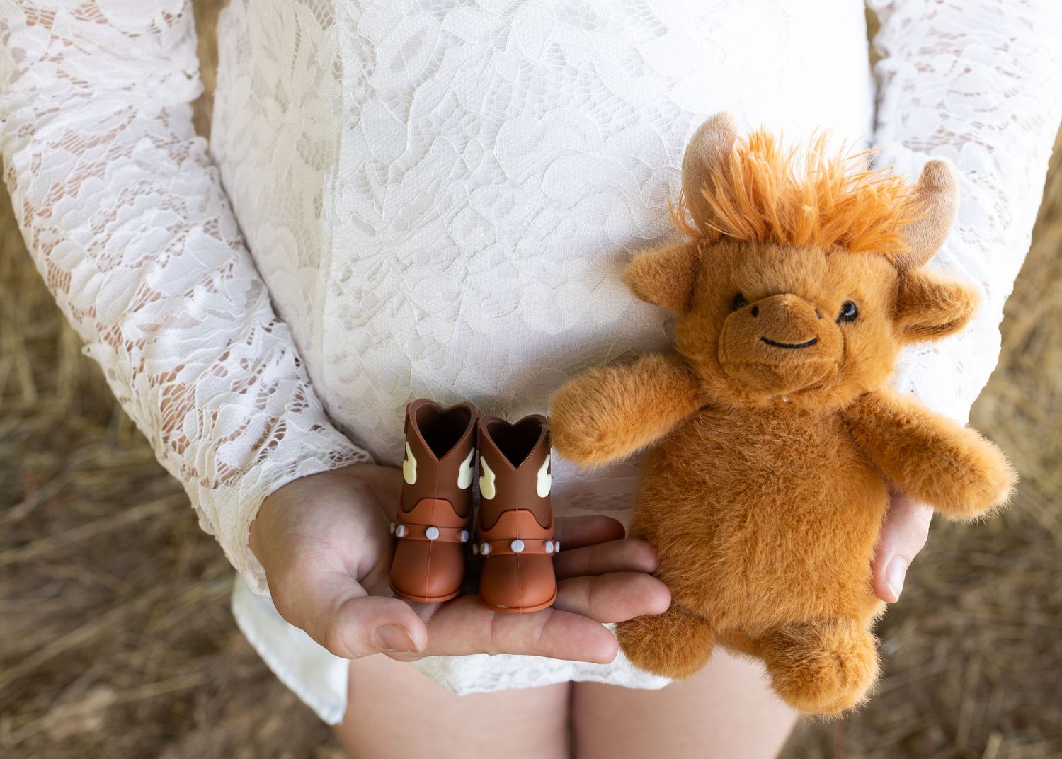 Pregnant woman holding tiny cowboy boots and a fluffy highland cow stuffed animal in her hands.
