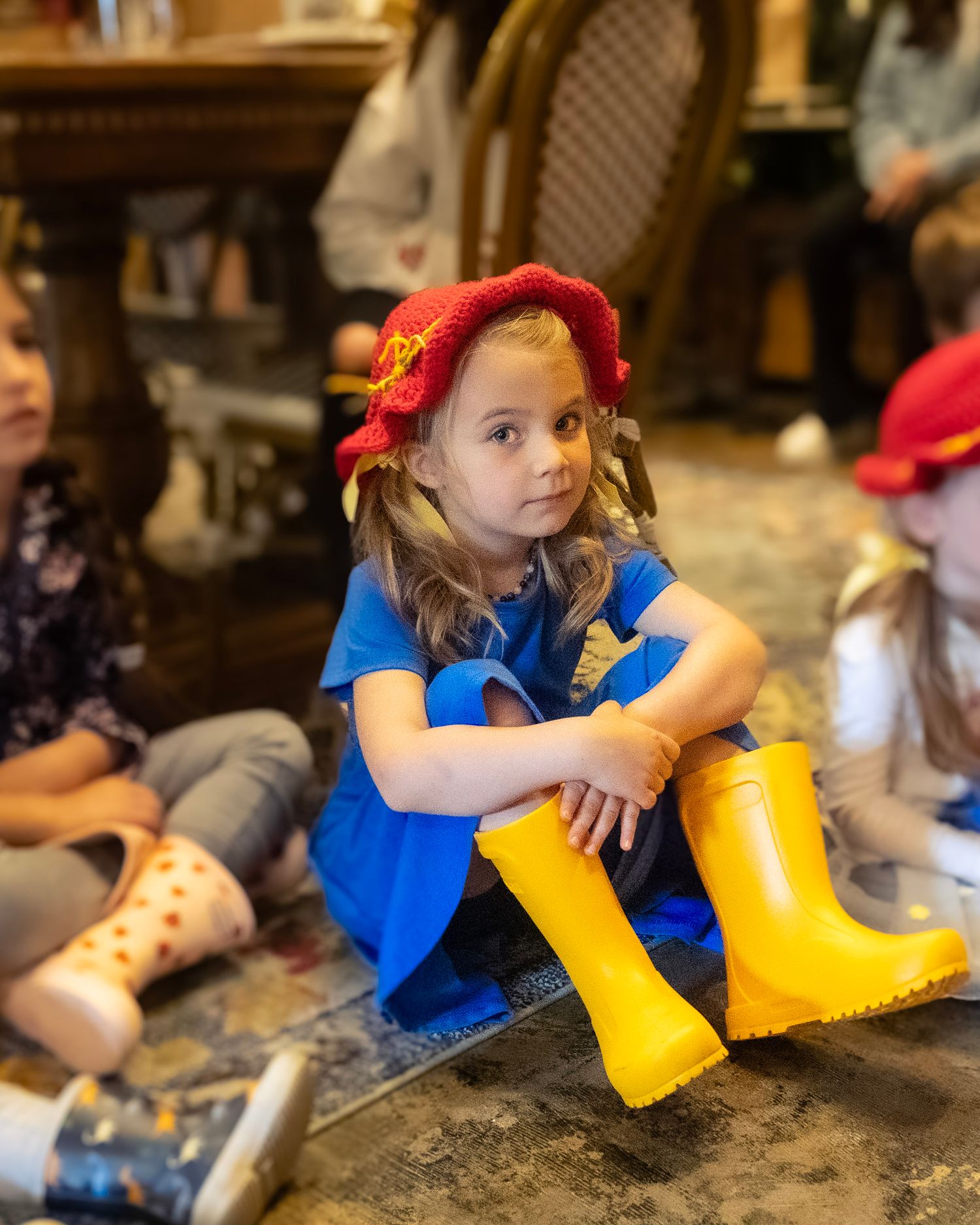Young girl in yellow rain boots, blue dress, and red knit hat sits cross-legged on floor among other children.
