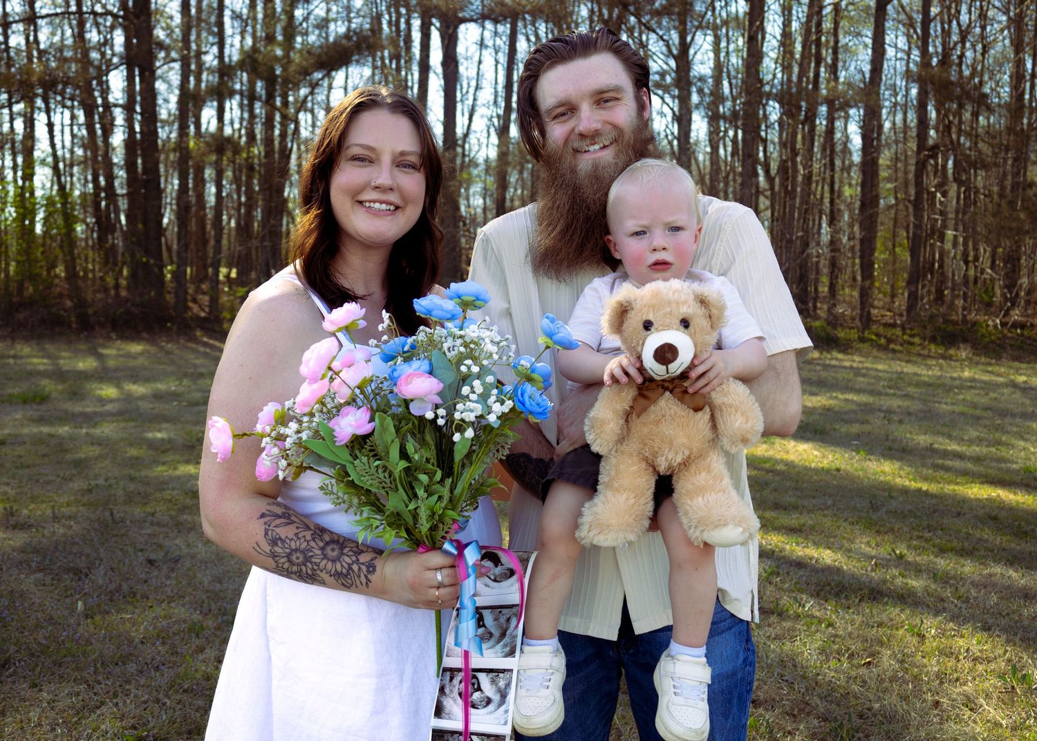 Family portrait of smiling couple with tattooed woman holding flowers and man carrying toddler with teddy bear outdoors.