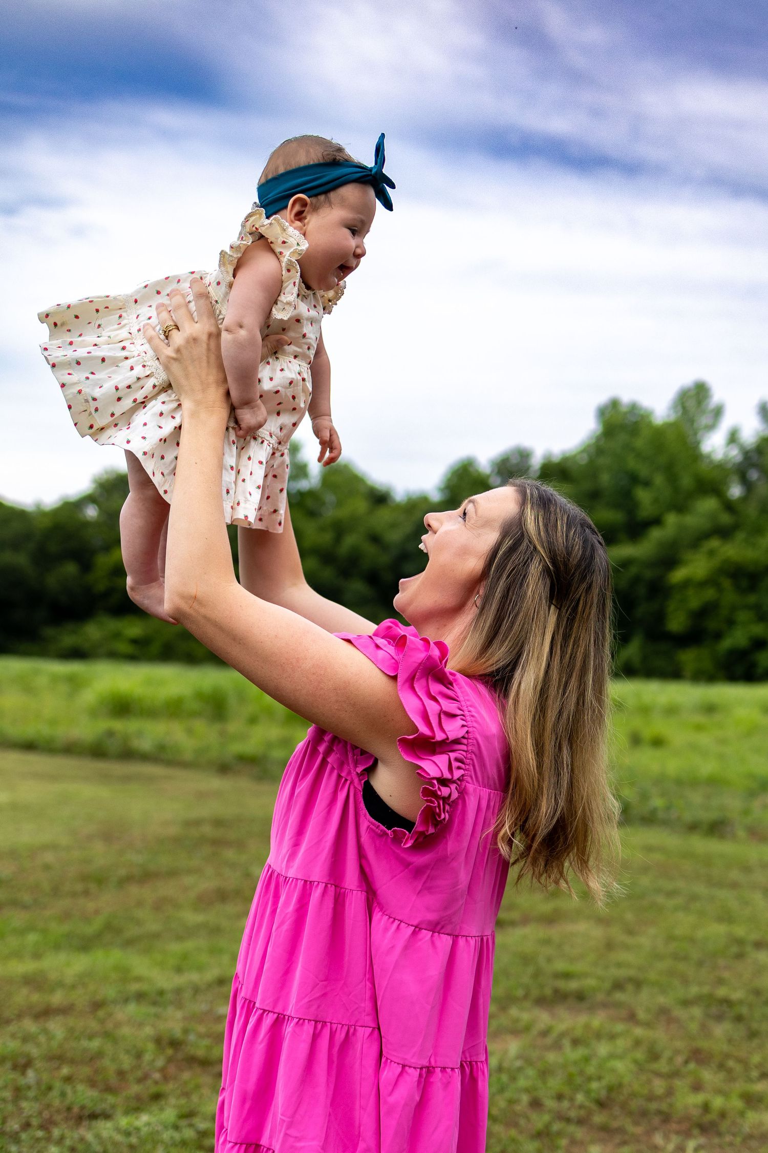 A mother in a pink dress joyfully lifts her baby girl in a polka dot dress outdoors in a green field.