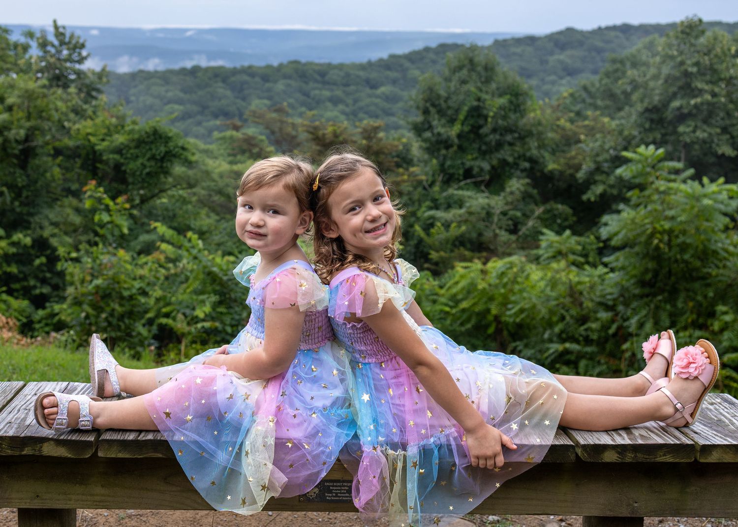 Two young girls in colorful fairy dresses sit back-to-back on a wooden bench with lush green hills in the background.