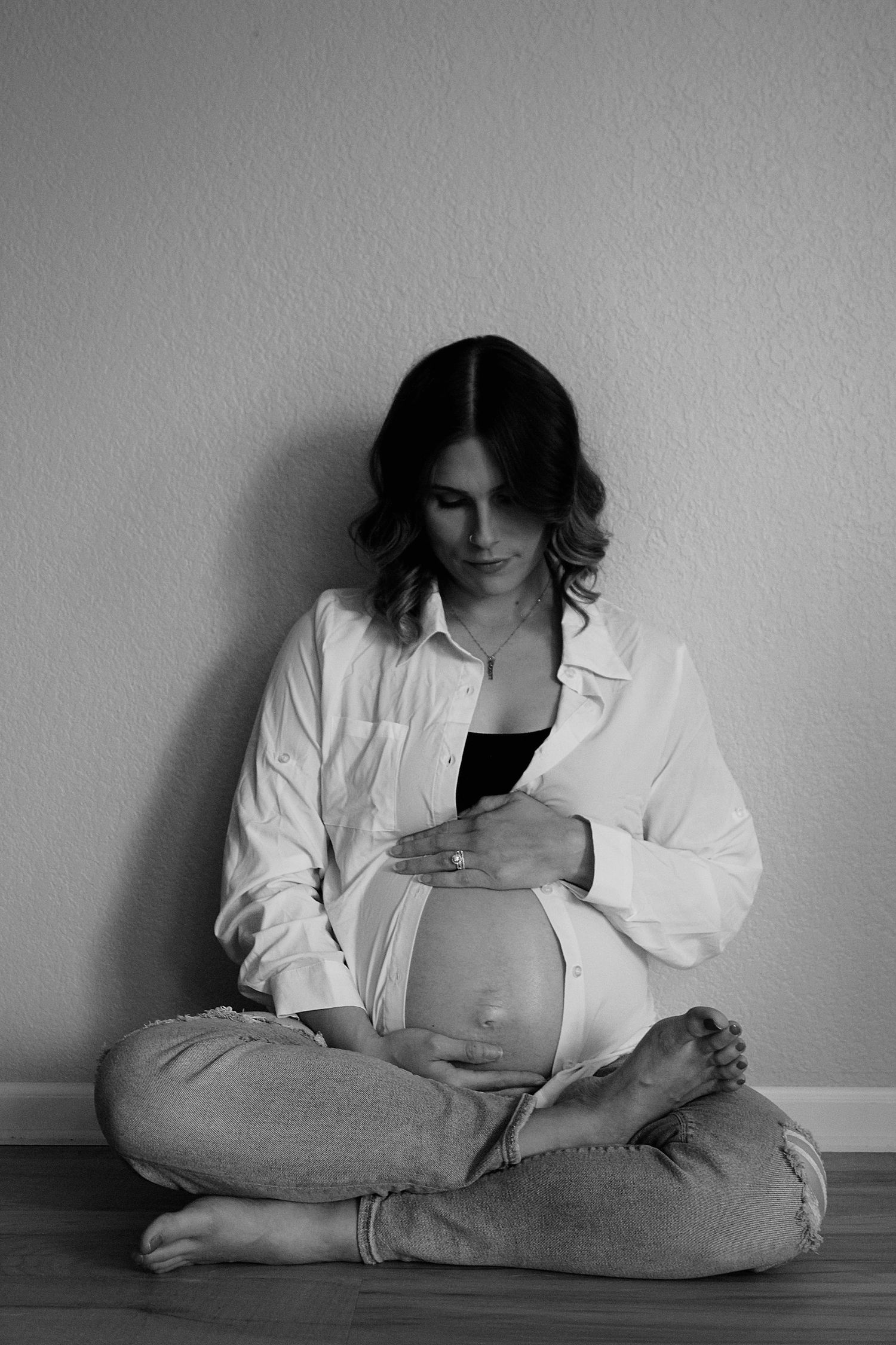 Black and white photo of a pregnant woman sitting cross-legged on floor, cradling her baby bump in a white shirt.