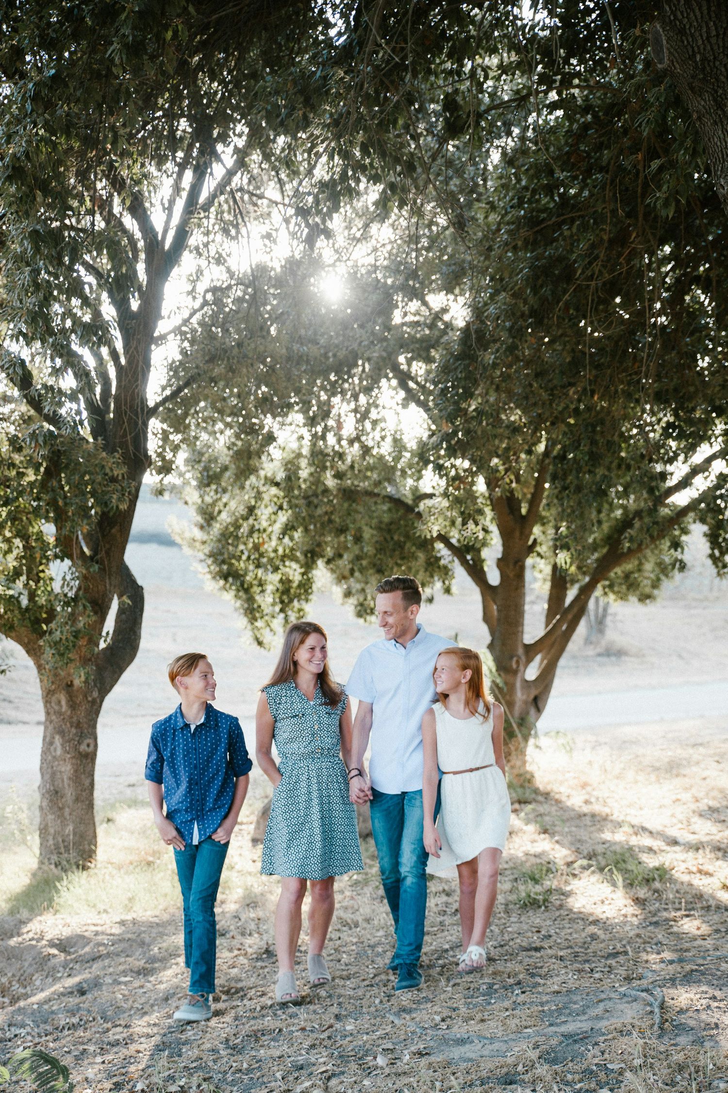 Family of four walking together under sunlit trees in a scenic outdoor setting, smiling and holding hands on a sunny day.