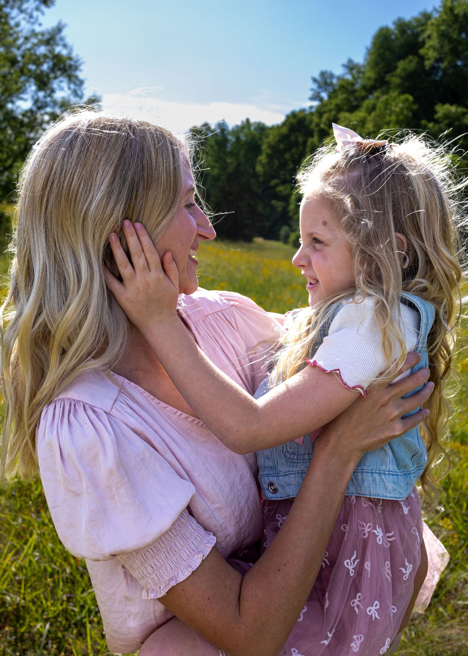 A blonde mother holds her young daughter in a sunny meadow, both smiling at each other in pink outfits.