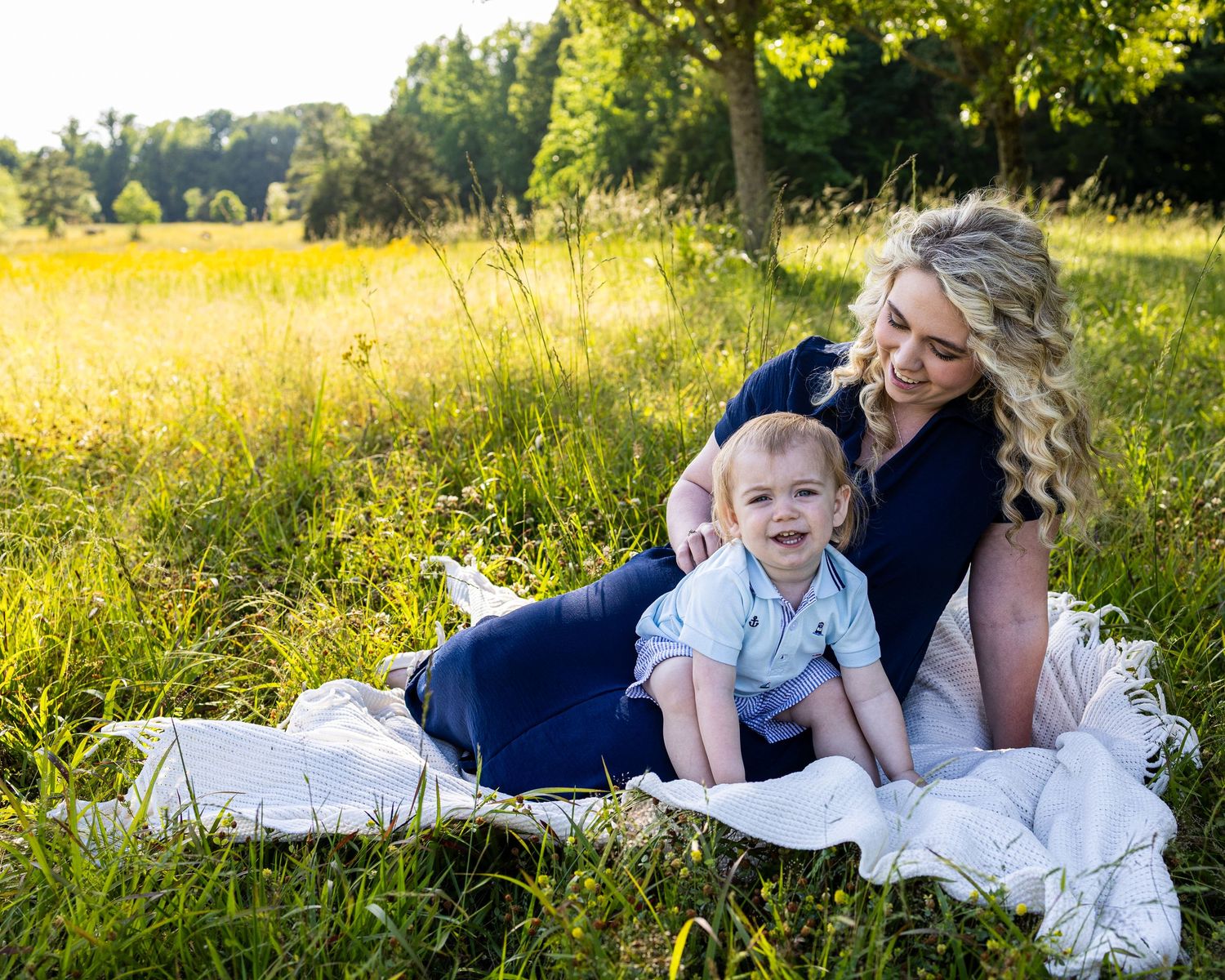 A smiling mother with curly blonde hair sits with her happy toddler boy on a white blanket in a sunny green meadow.