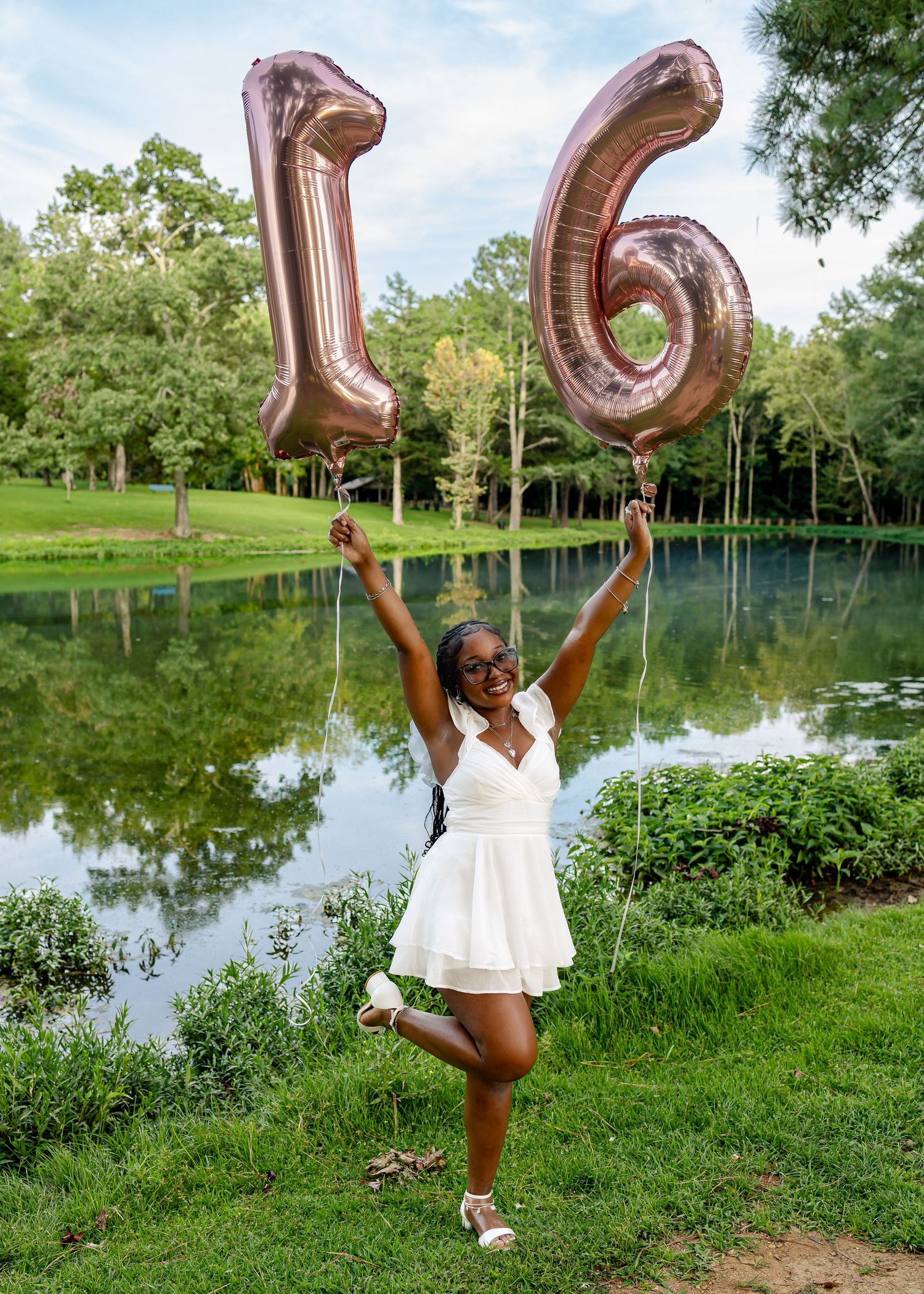 A joyful young woman in a white dress holds rose gold number 16 balloons outdoors by a pond for her sweet sixteen.