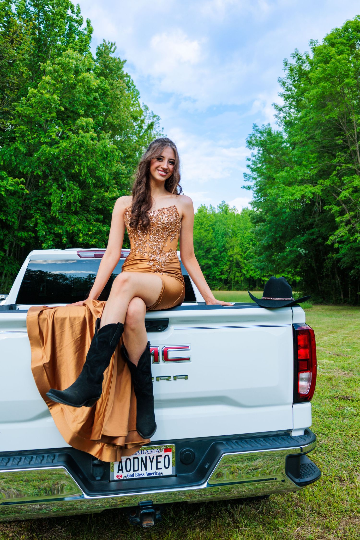Young woman in gold sequin gown and cowboy boots sitting on tailgate of white GMC truck outdoors.