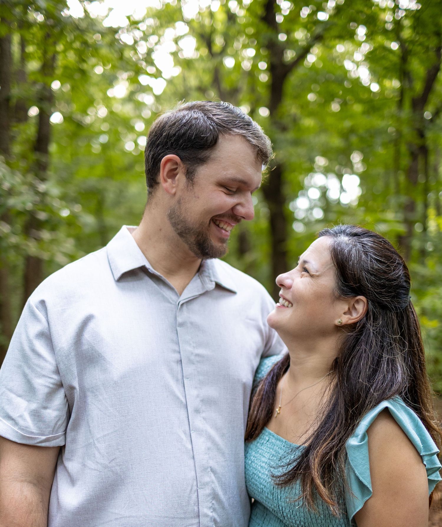 A smiling couple gazes at each other outdoors in a lush green forest setting, man in blue shirt, woman in teal top.