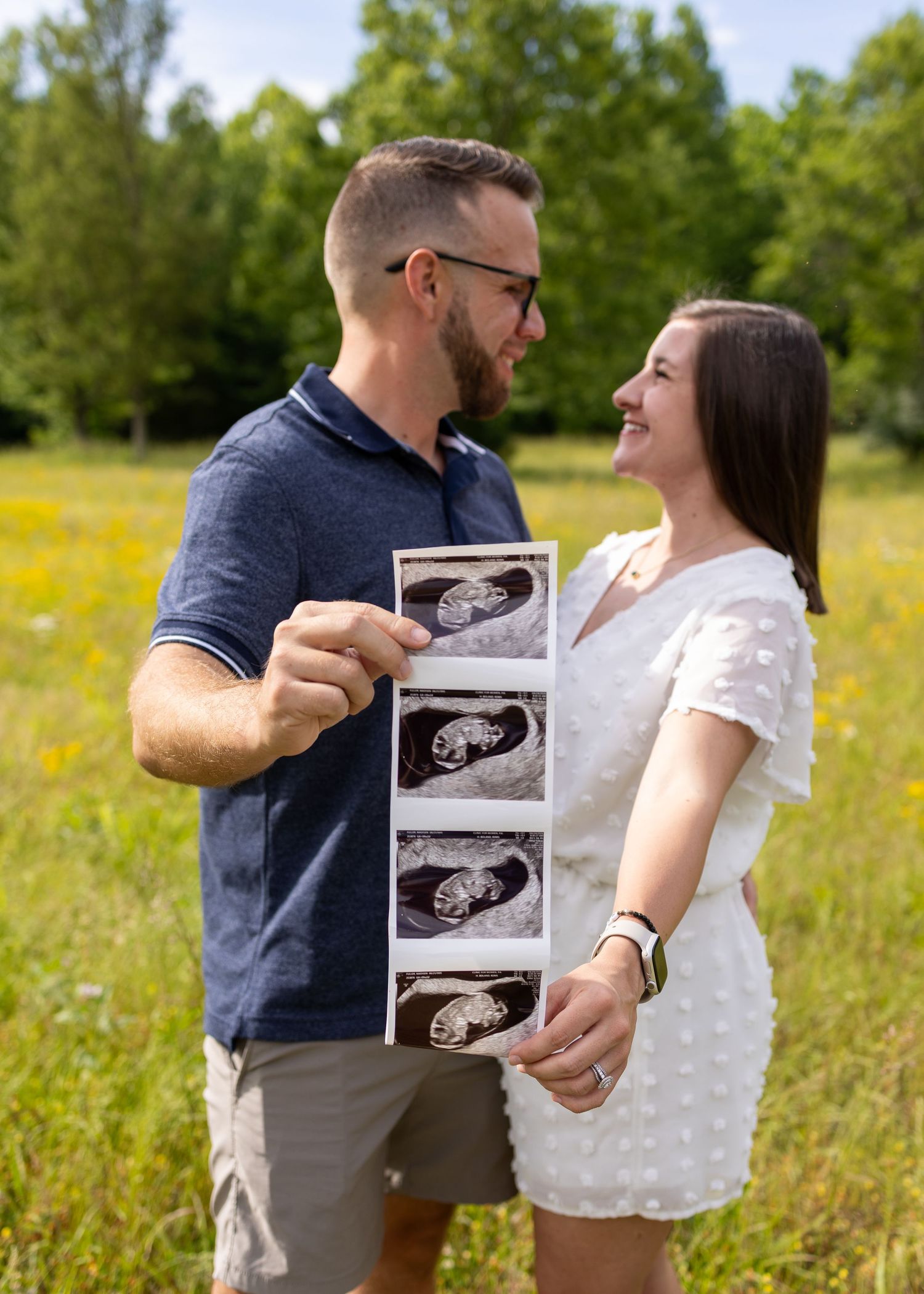 Happy couple holding ultrasound photos in a sunny meadow, announcing their pregnancy outdoors.