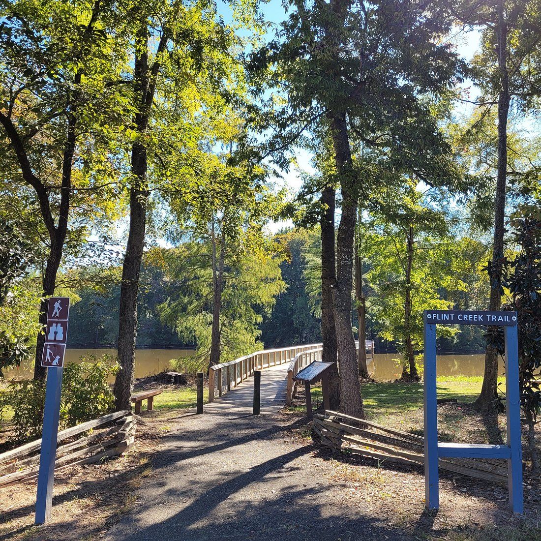 Flint Creek Trail entrance with blue signs, wooden bridge, and sunlit trees near a calm lake in a scenic park.