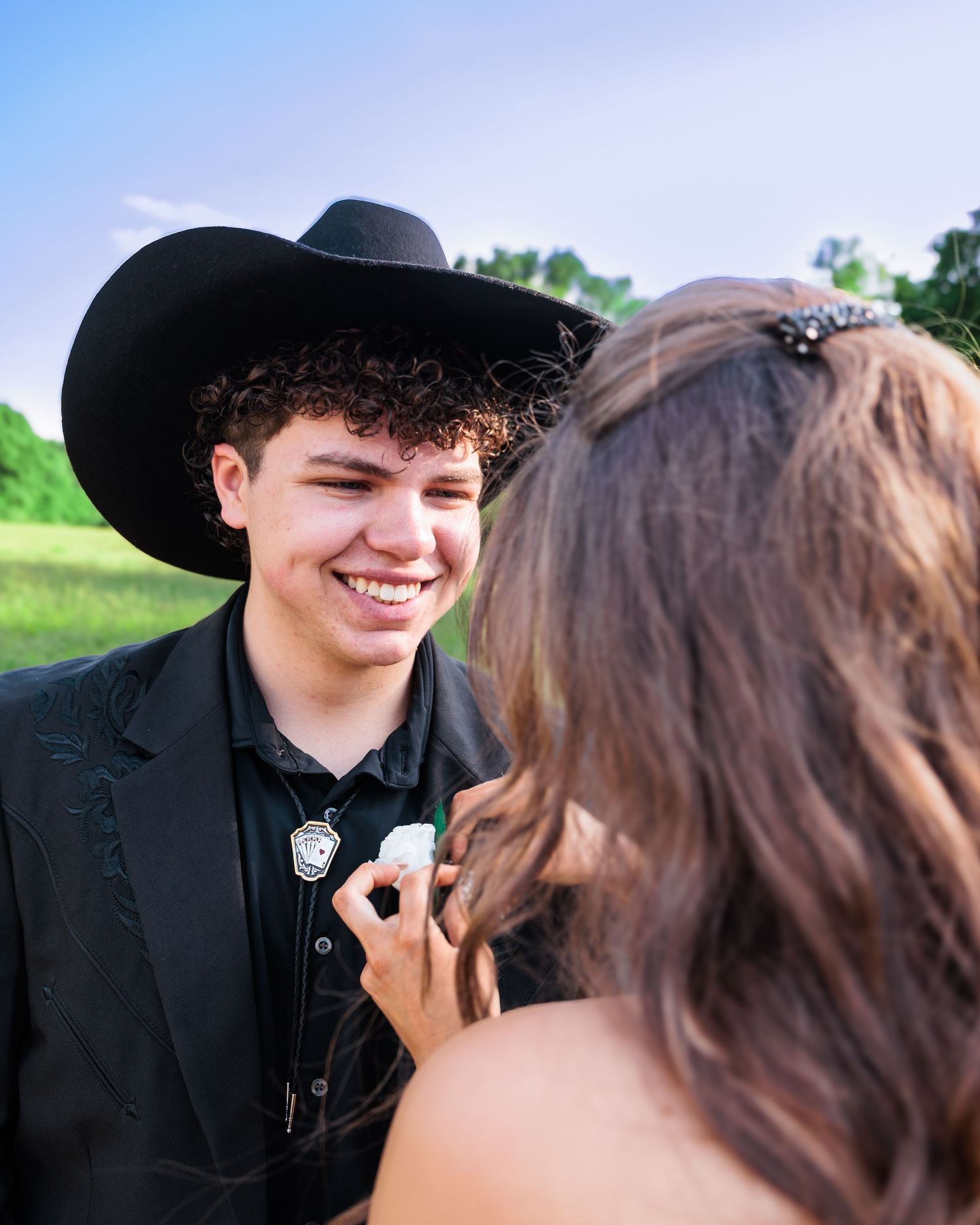A smiling young man in a black cowboy hat and suit receives a boutonniere at a prom or formal event outdoors.