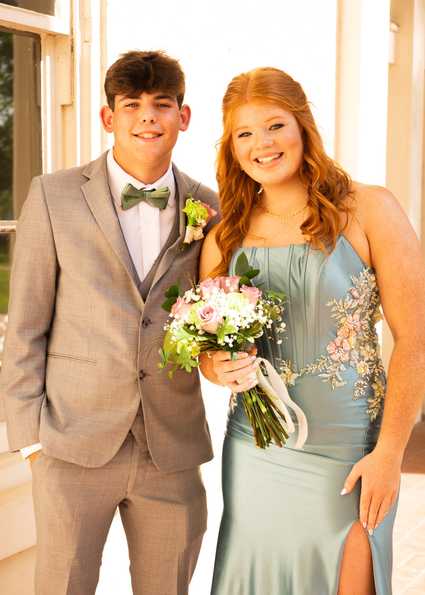Young couple dressed for prom, boy in gray suit with bow tie, girl in sage green floral gown holding bouquet.