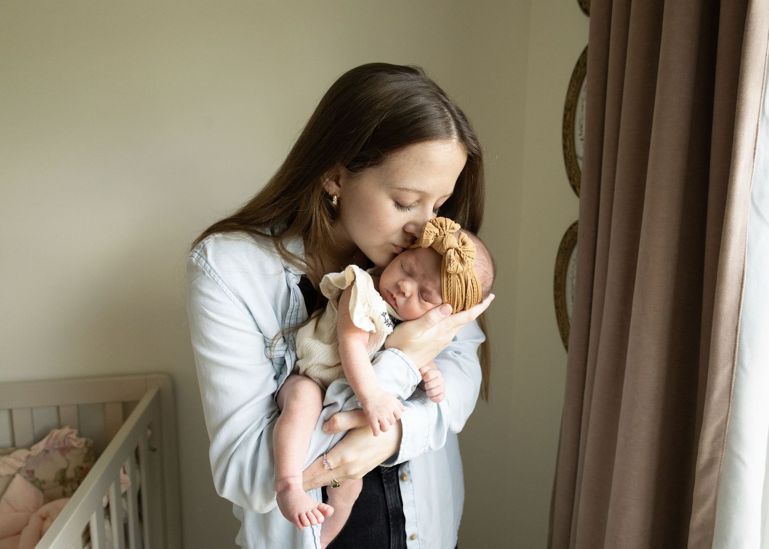 A mother tenderly kisses her newborn baby while standing in a softly lit nursery room.