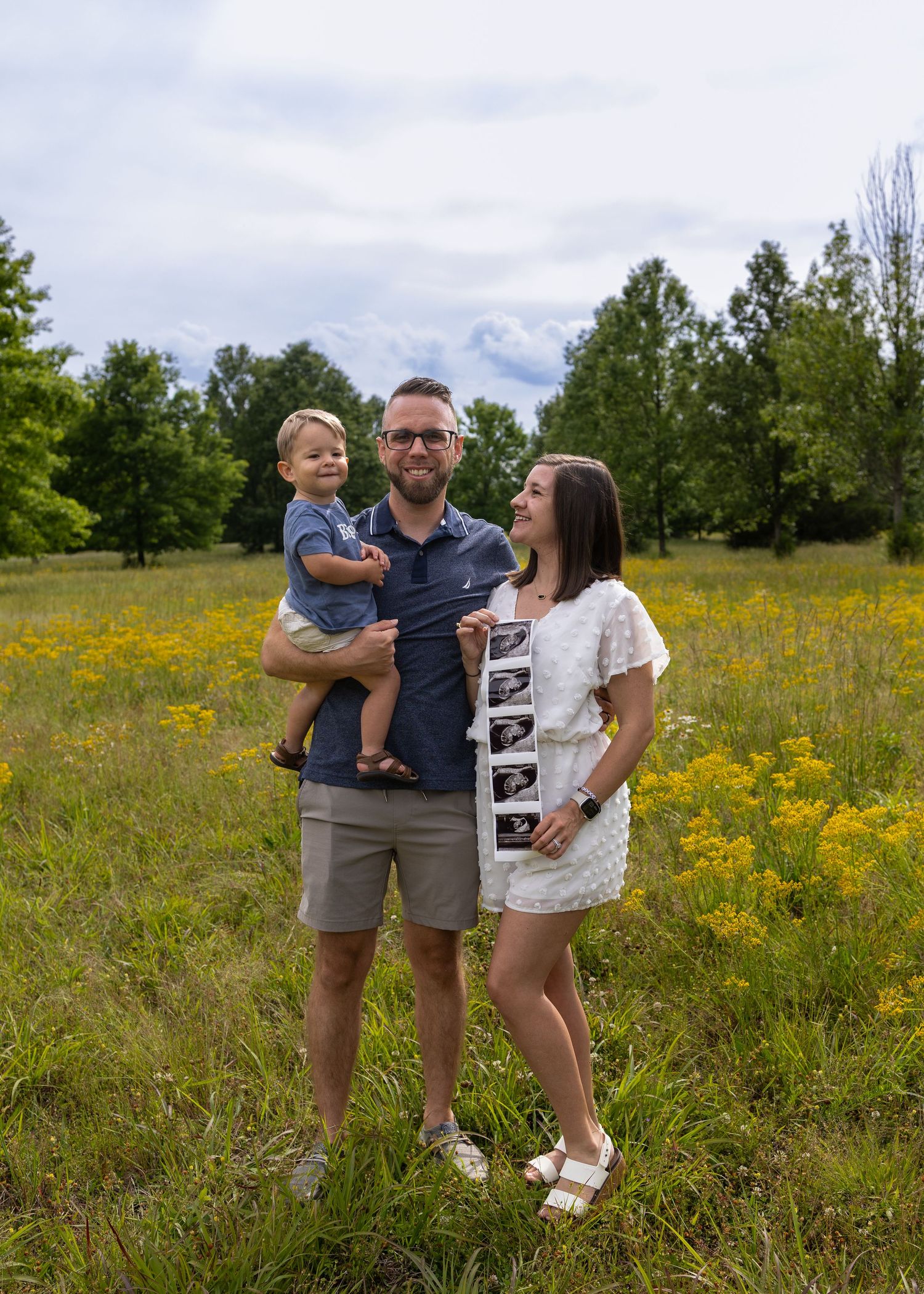 A happy family of three poses in a sunny meadow, with a man holding a toddler and a woman displaying ultrasound photos.