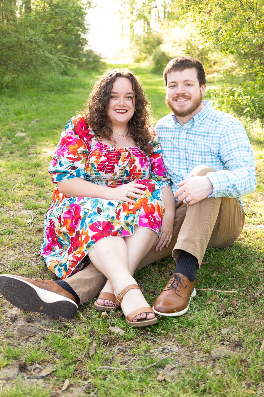 Smiling pregnant couple sitting on grass in a sunny park, woman in floral dress, man in plaid shirt and khakis.