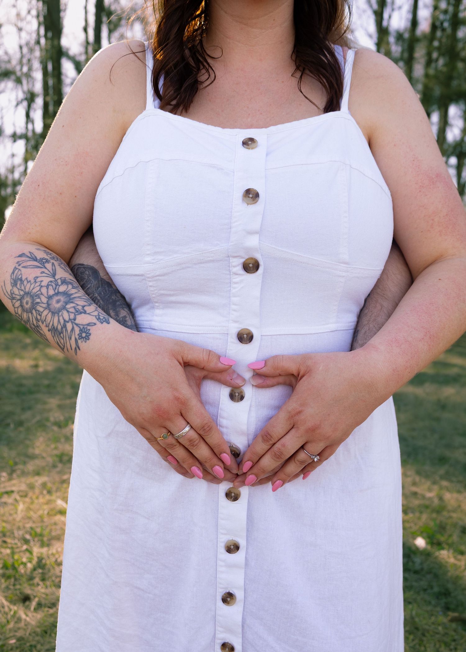 Woman in white button-down sundress with floral arm tattoo forming heart shape with hands outdoors.
