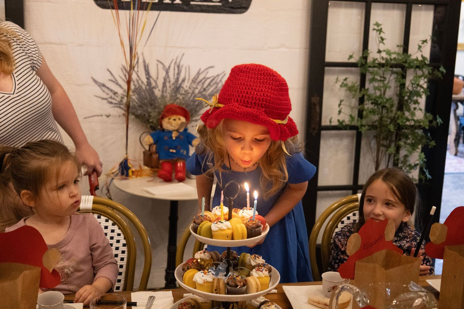 Girl in red hat blowing out birthday candles on tiered cake stand, with children watching at a party table.