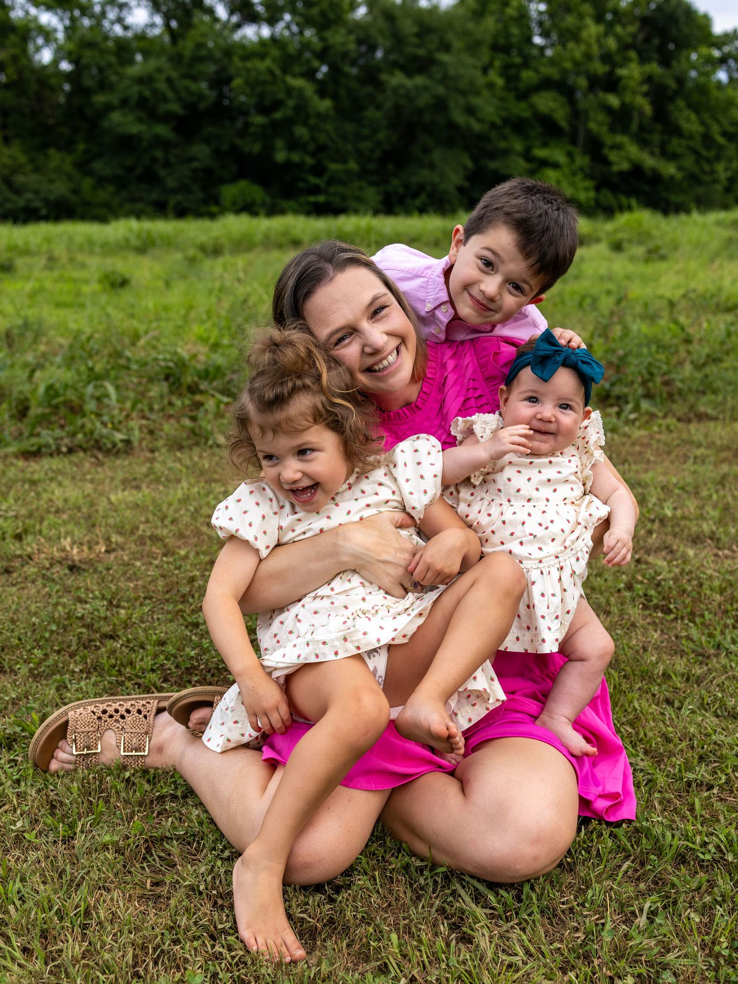 A smiling mother in a pink dress sits on grass holding three young children, including a baby with a blue bow.