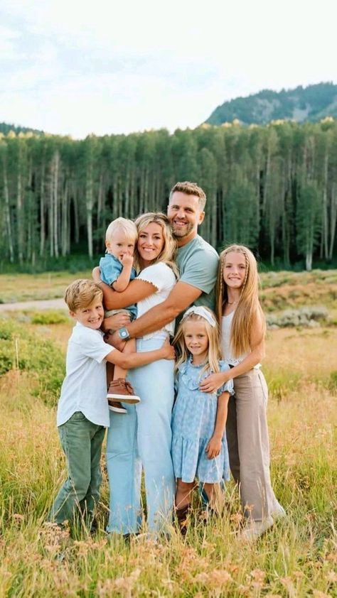 Happy family of six posing in a sunny meadow with tall green trees and mountains in the background.