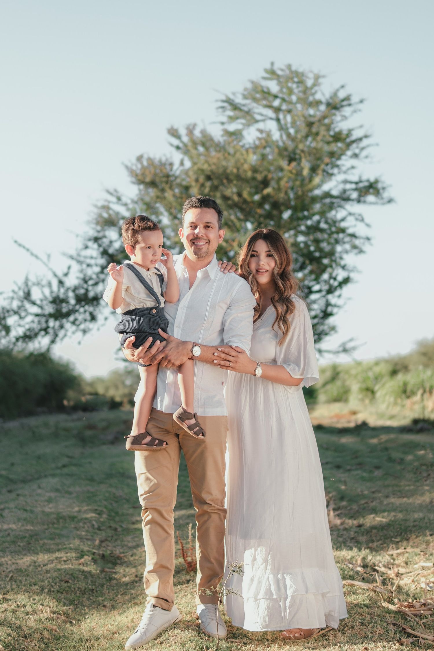 A happy young family with a toddler poses outdoors in neutral tones against a natural green backdrop.