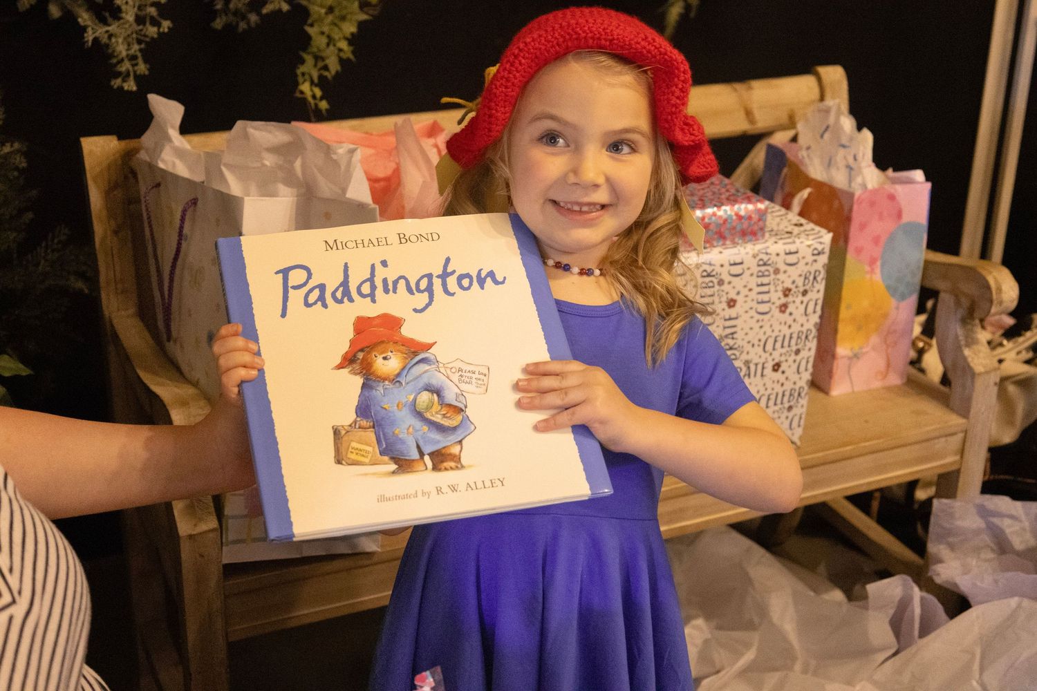 Young girl in red hat and blue dress proudly holds a Paddington Bear book while sitting on a wooden bench.