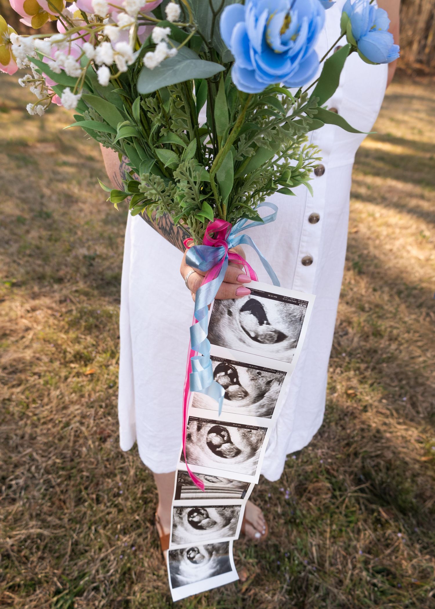 Pregnant woman in white dress holding flower bouquet with ultrasound photos and pink blue ribbons for gender reveal.