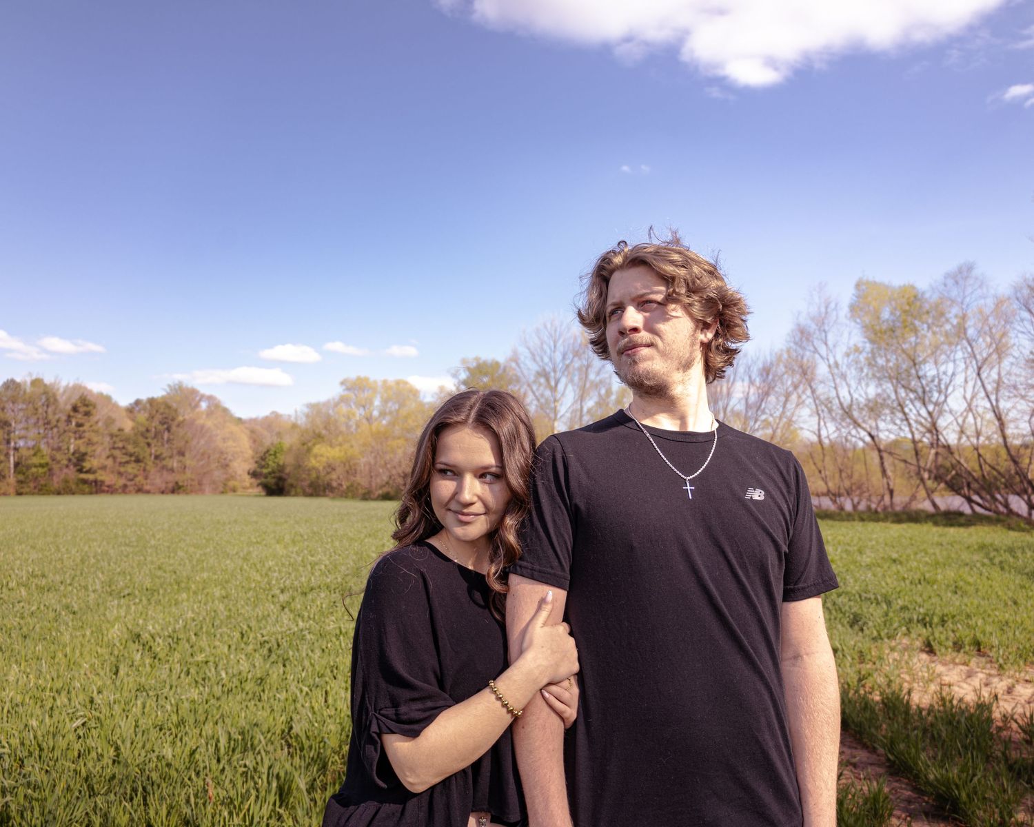 Two teenagers in black outfits posing together in a green field under a bright blue sky on a sunny day.
