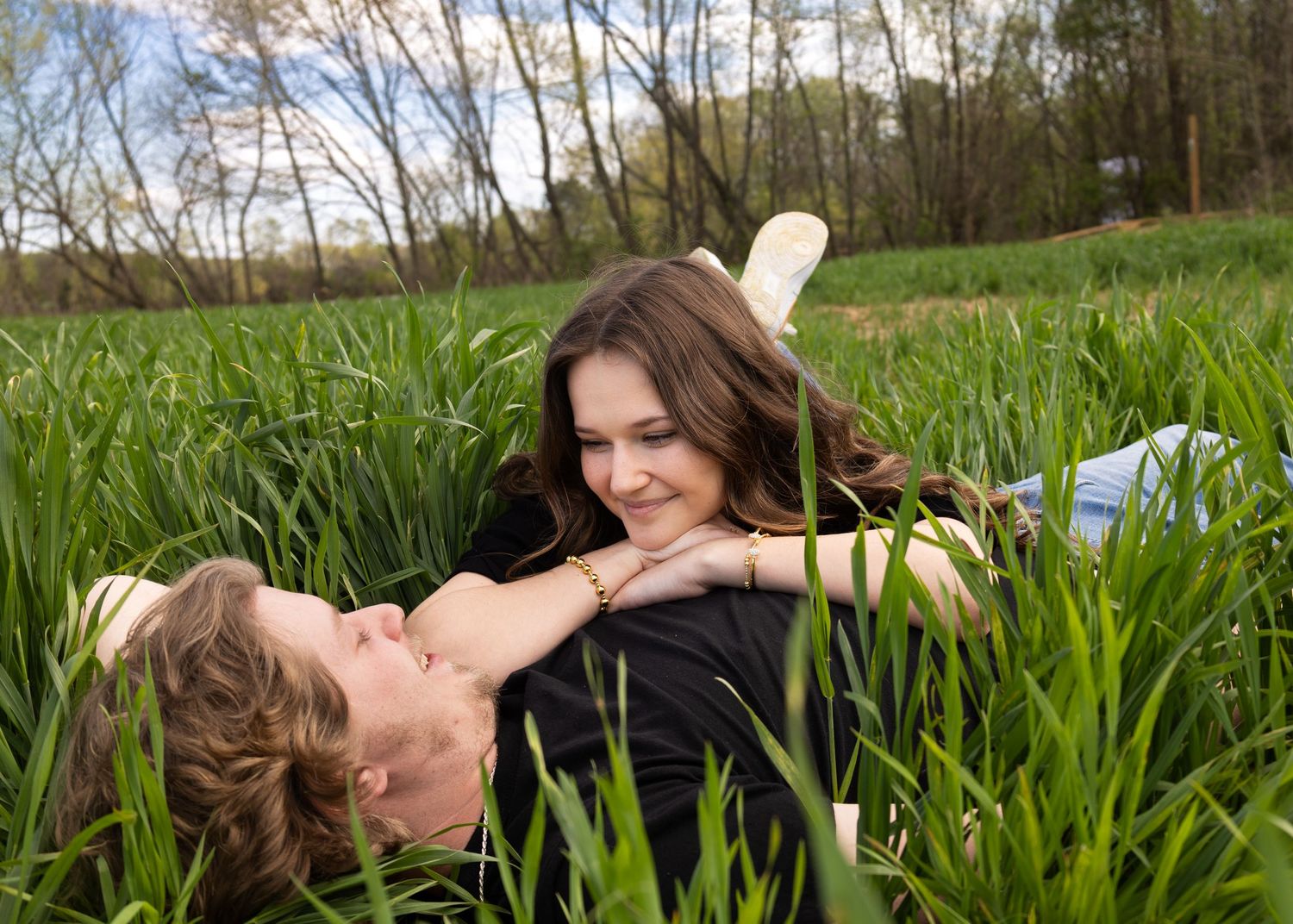 Young couple lying in tall green grass, smiling and gazing at each other on a sunny day outdoors.