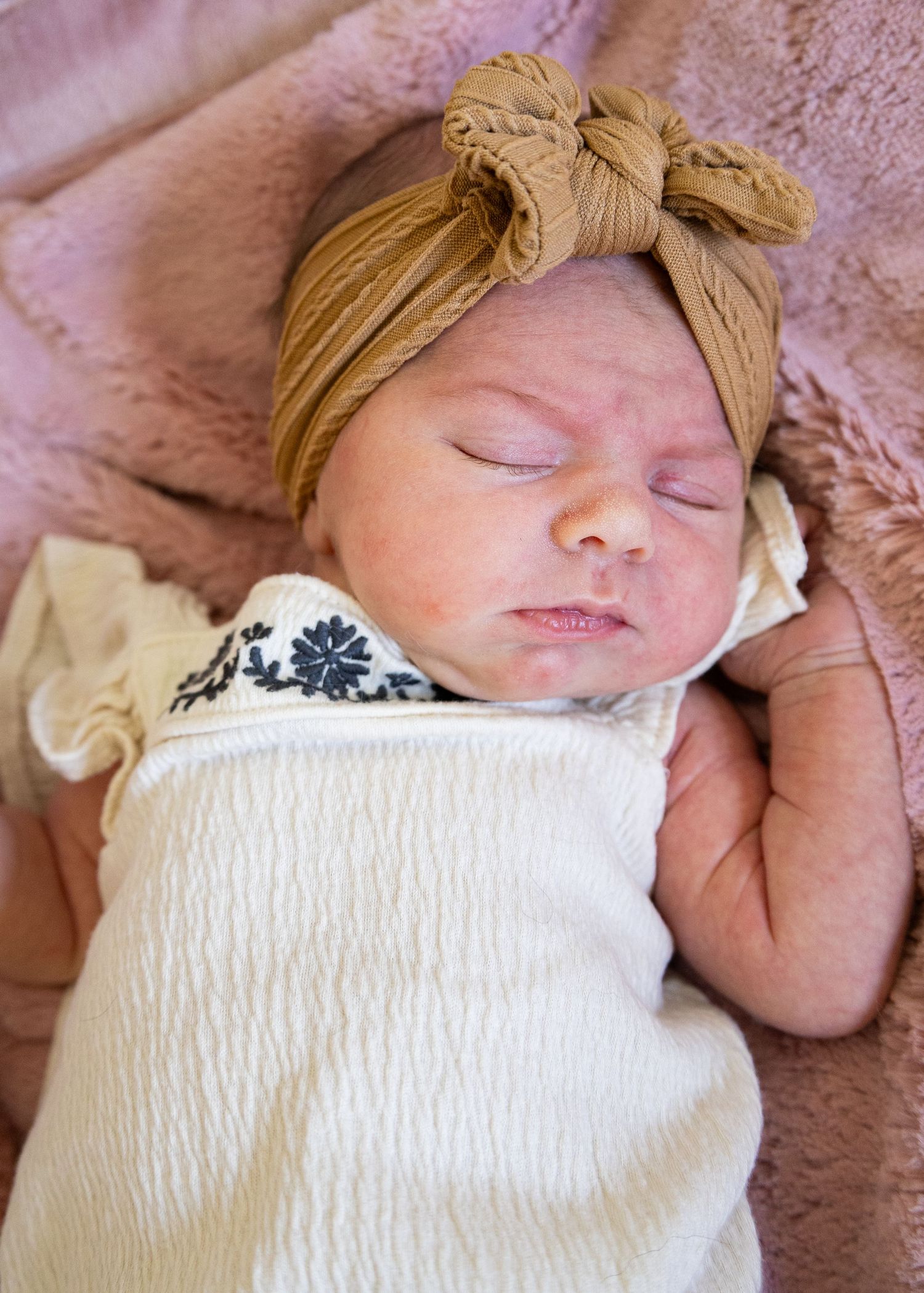 Sleeping newborn baby girl wearing a golden bow headband, swaddled in a white blanket on a pink background.