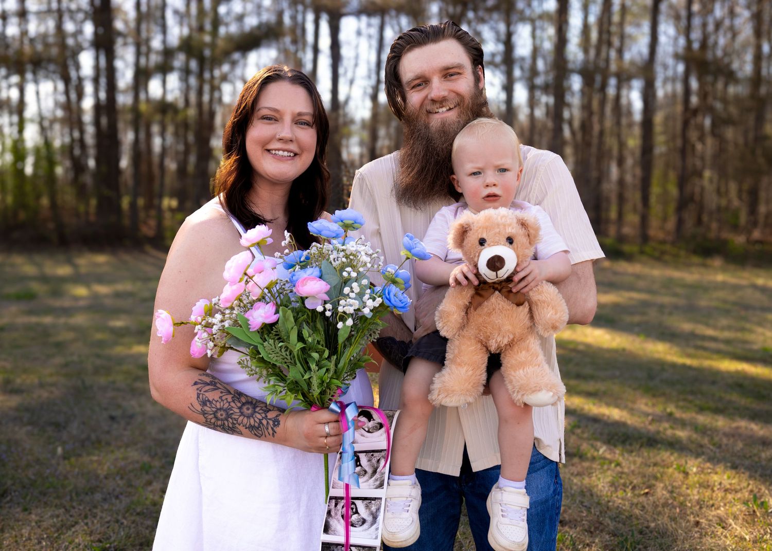 A smiling couple stands outdoors holding flowers and a teddy bear with a young toddler, surrounded by trees.