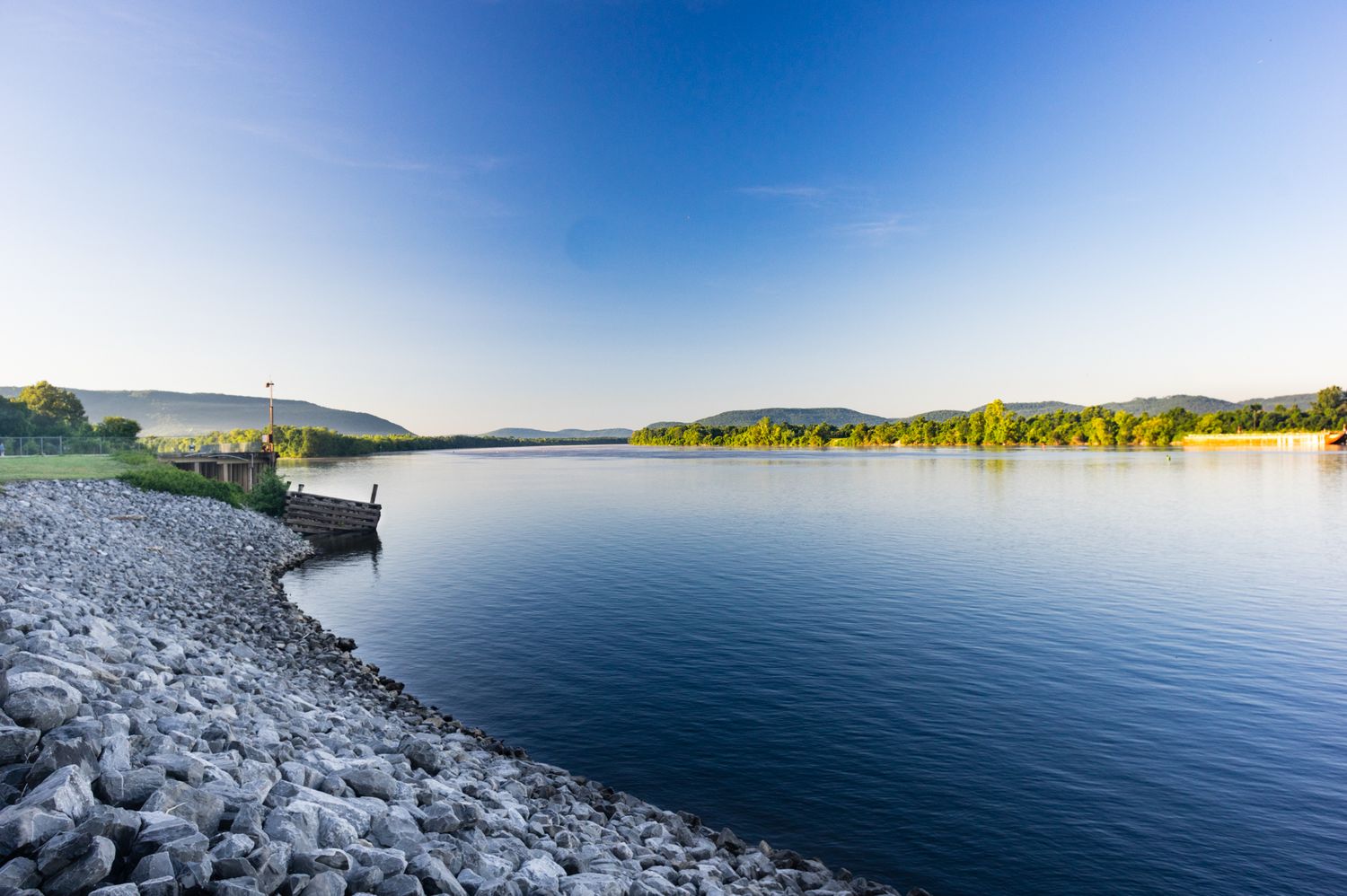 Serene lake with rocky shoreline, calm blue water, lush green trees, and rolling hills under a clear blue sky.