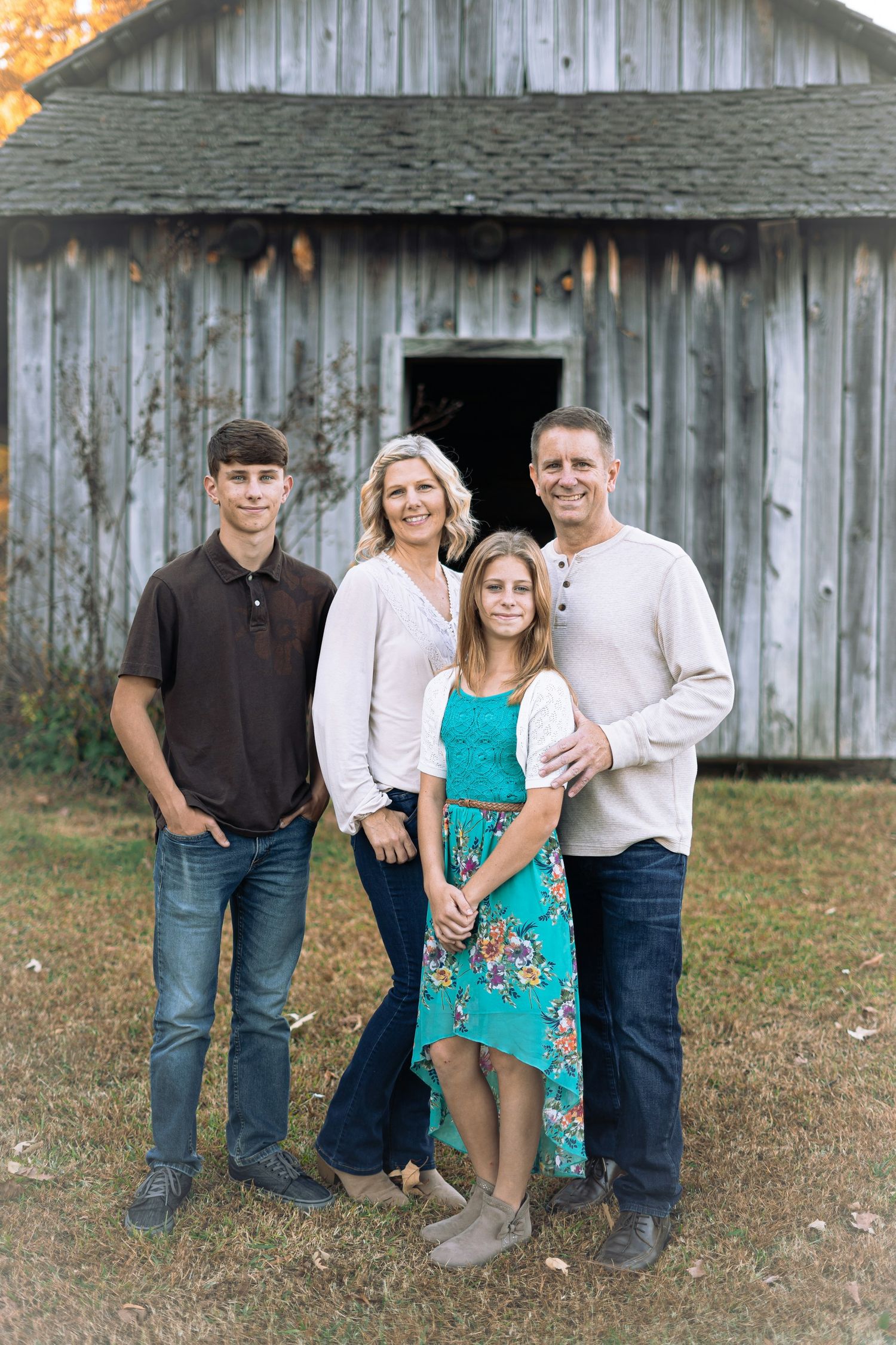 Family of four smiling together in front of a rustic wooden barn during autumn outdoor photo session.