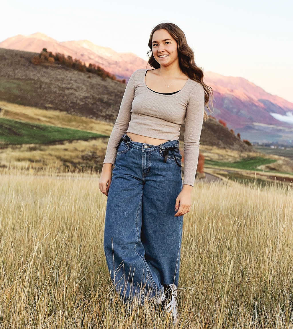 Young woman in beige long-sleeve top and wide-leg jeans smiling in a mountain meadow at sunset.
