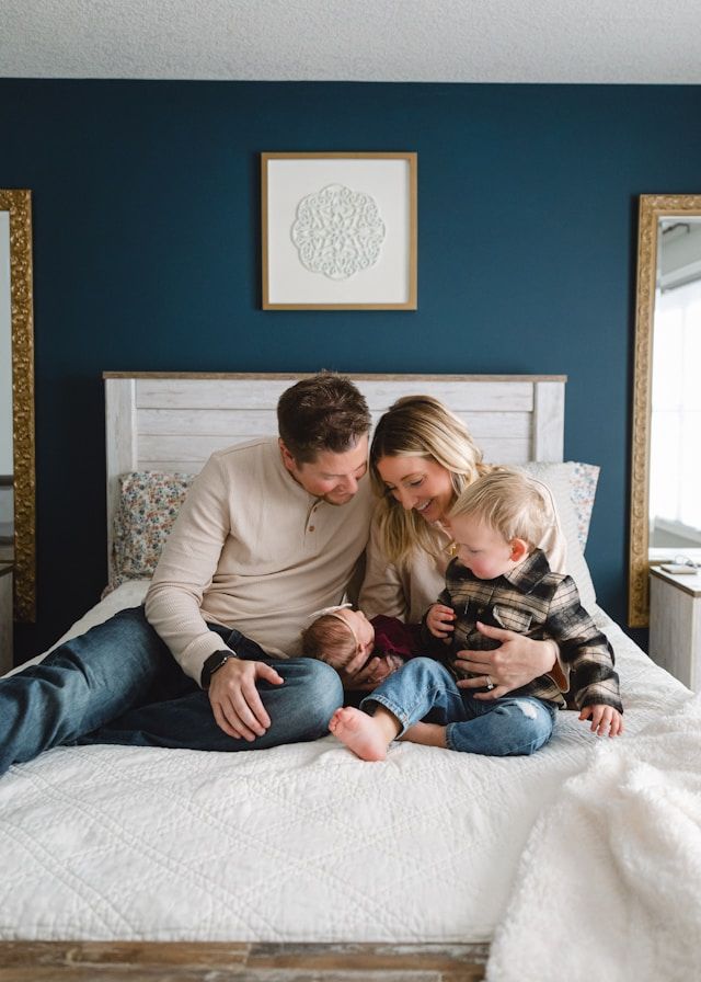 Family of four sitting on a white bed against a navy blue wall, parents admiring their newborn baby and toddler.