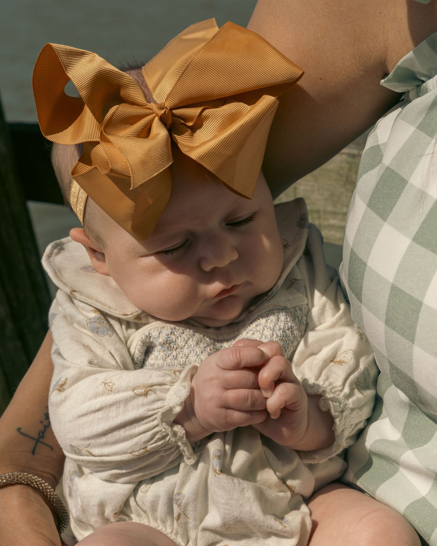 Baby in a cream-colored dress with a large matching bow headband looks down while sitting on a checkered blanket.