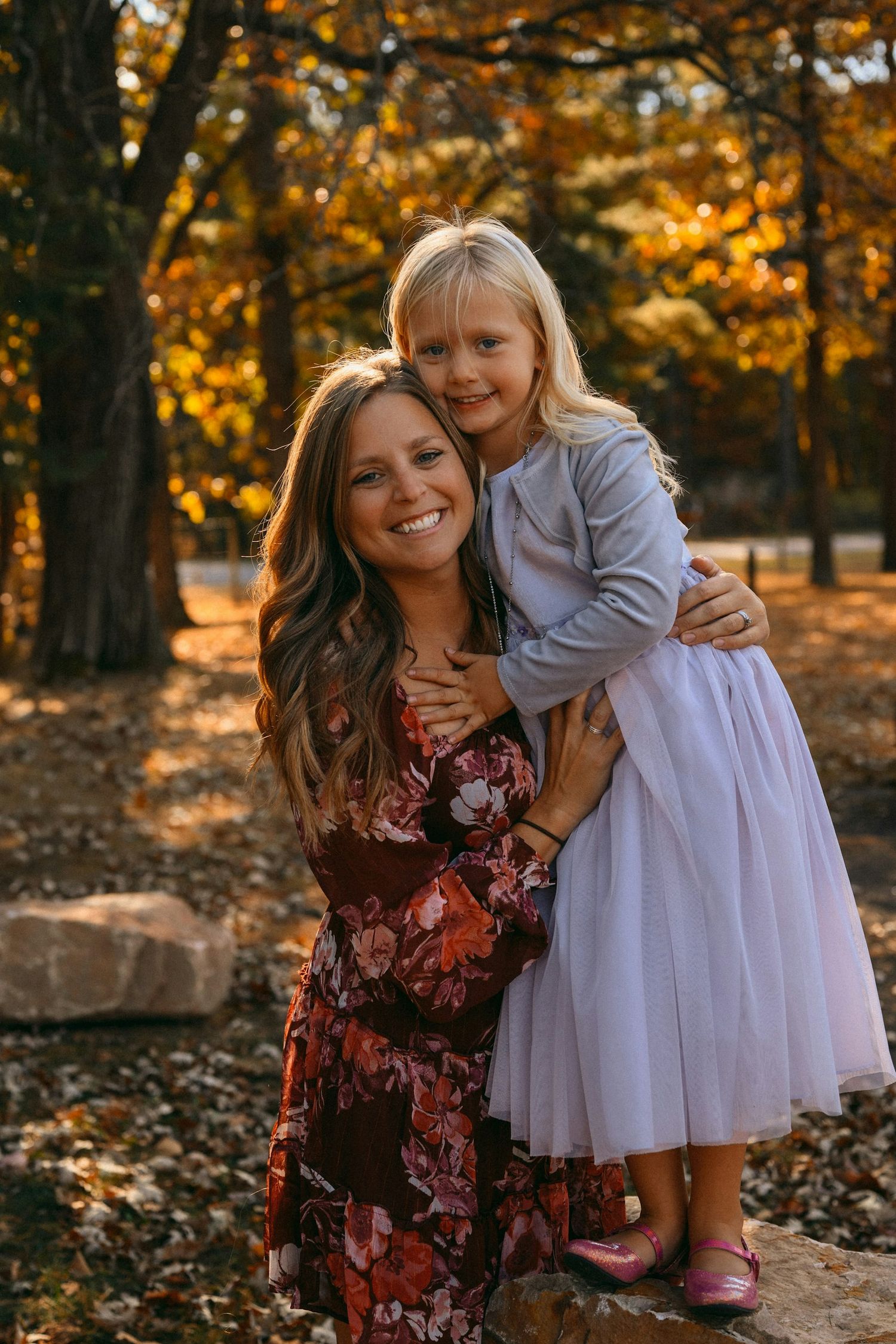A smiling mother in a floral dress hugs her blonde daughter in a tulle skirt outdoors among fall foliage.