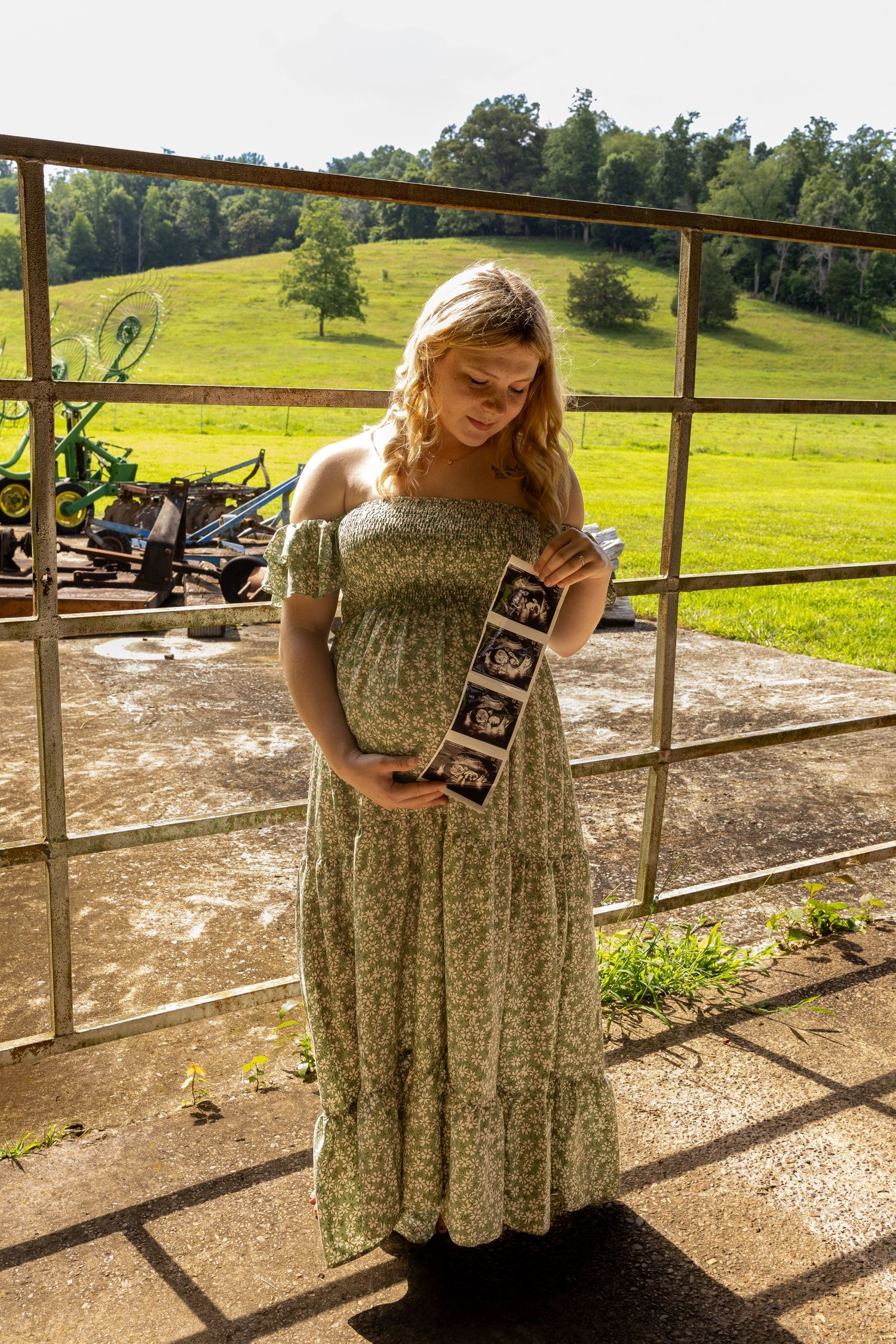 Pregnant woman in floral dress holding ultrasound photos outdoors on a sunny farm with green hills in background.