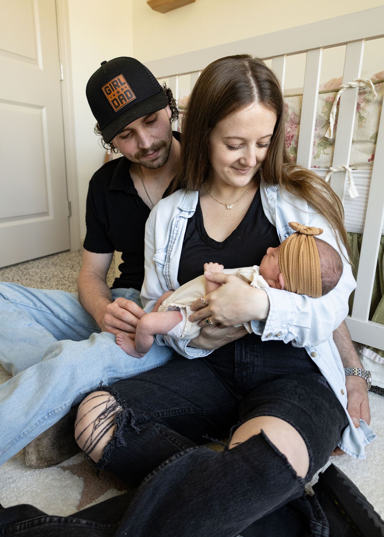 Young couple sitting on floor smiling at their newborn baby girl, man wearing a Girl Dad hat.