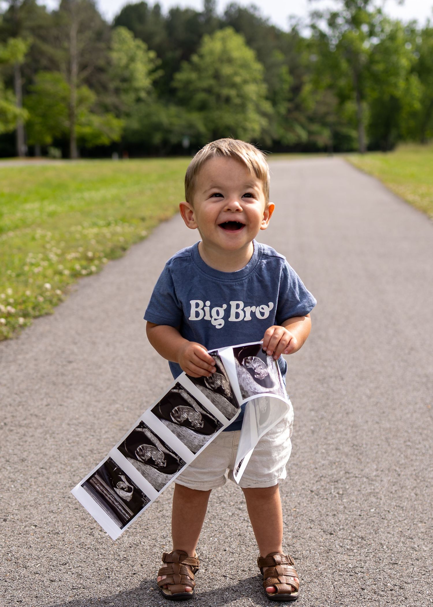 Smiling toddler in Big Bro shirt holding ultrasound photos on a park path, announcing pregnancy.