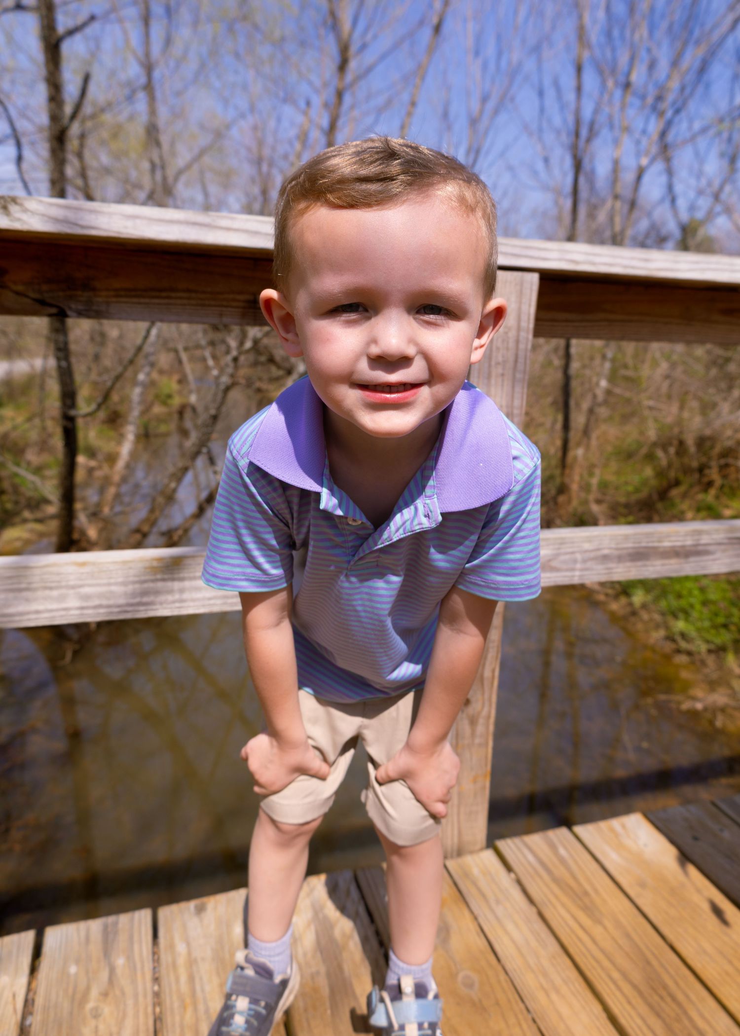 A young boy in a purple striped polo and khaki shorts smiles on a wooden bridge outdoors.